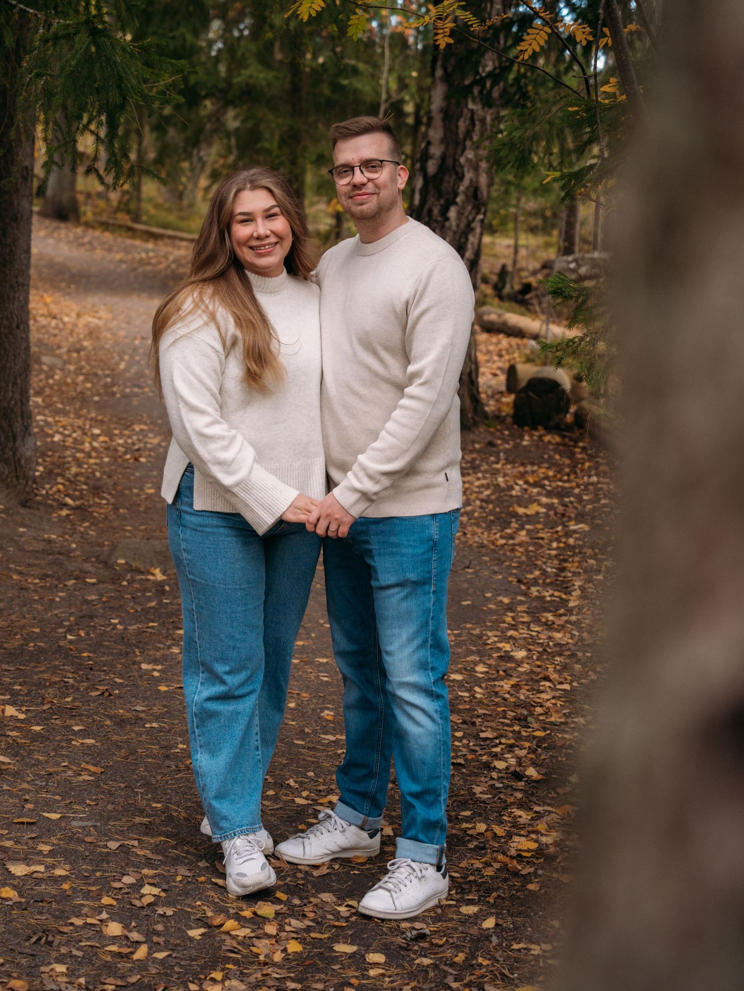 A couple in matching cream sweaters and blue jeans holds hands on a leaf-covered forest path in autumn.