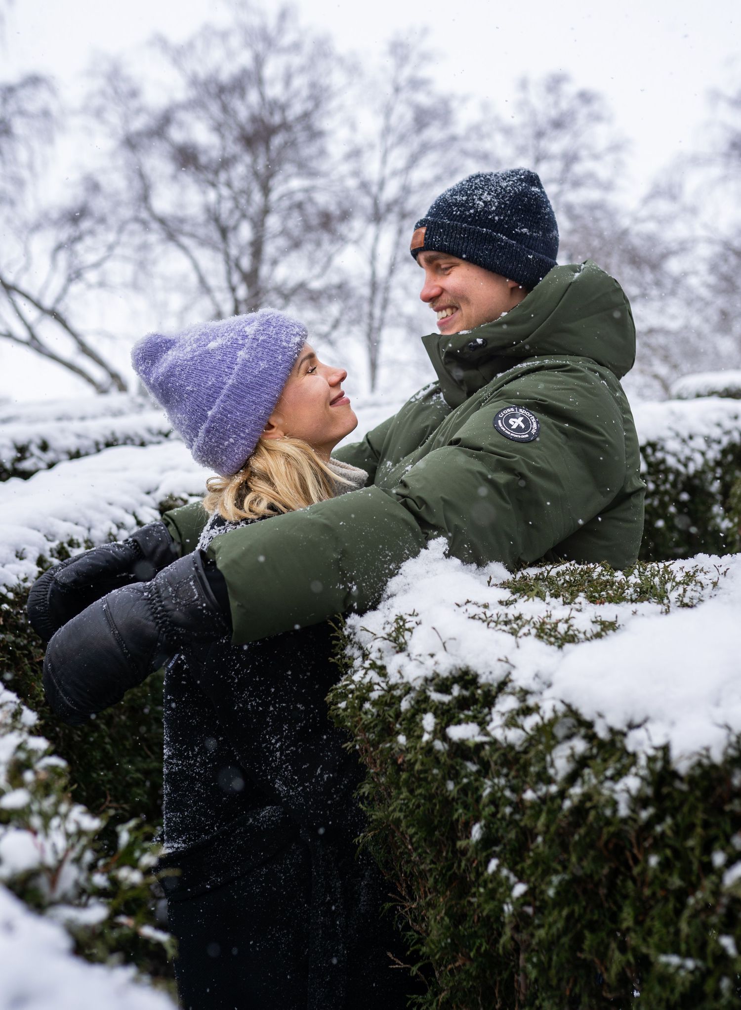 Couple embracing and smiling at each other outdoors in a snowy winter park, wearing colorful knit hats and warm jackets.