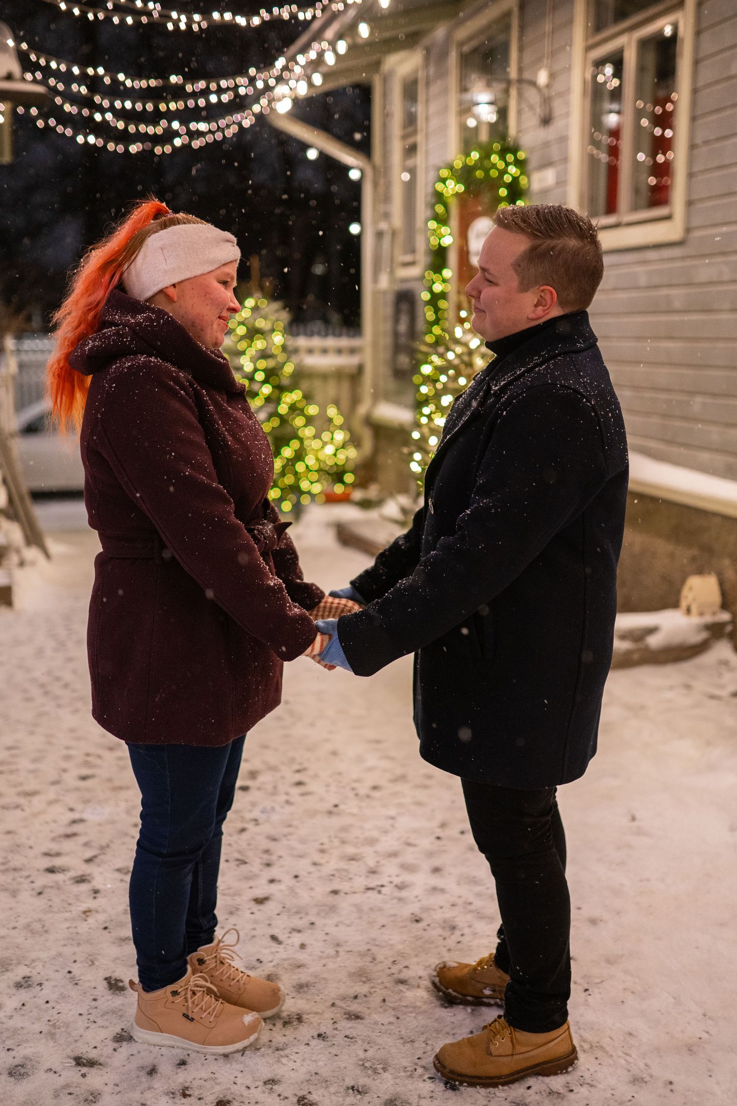 Couple holding hands in snowy winter night, Christmas lights and decorated tree glowing behind them.