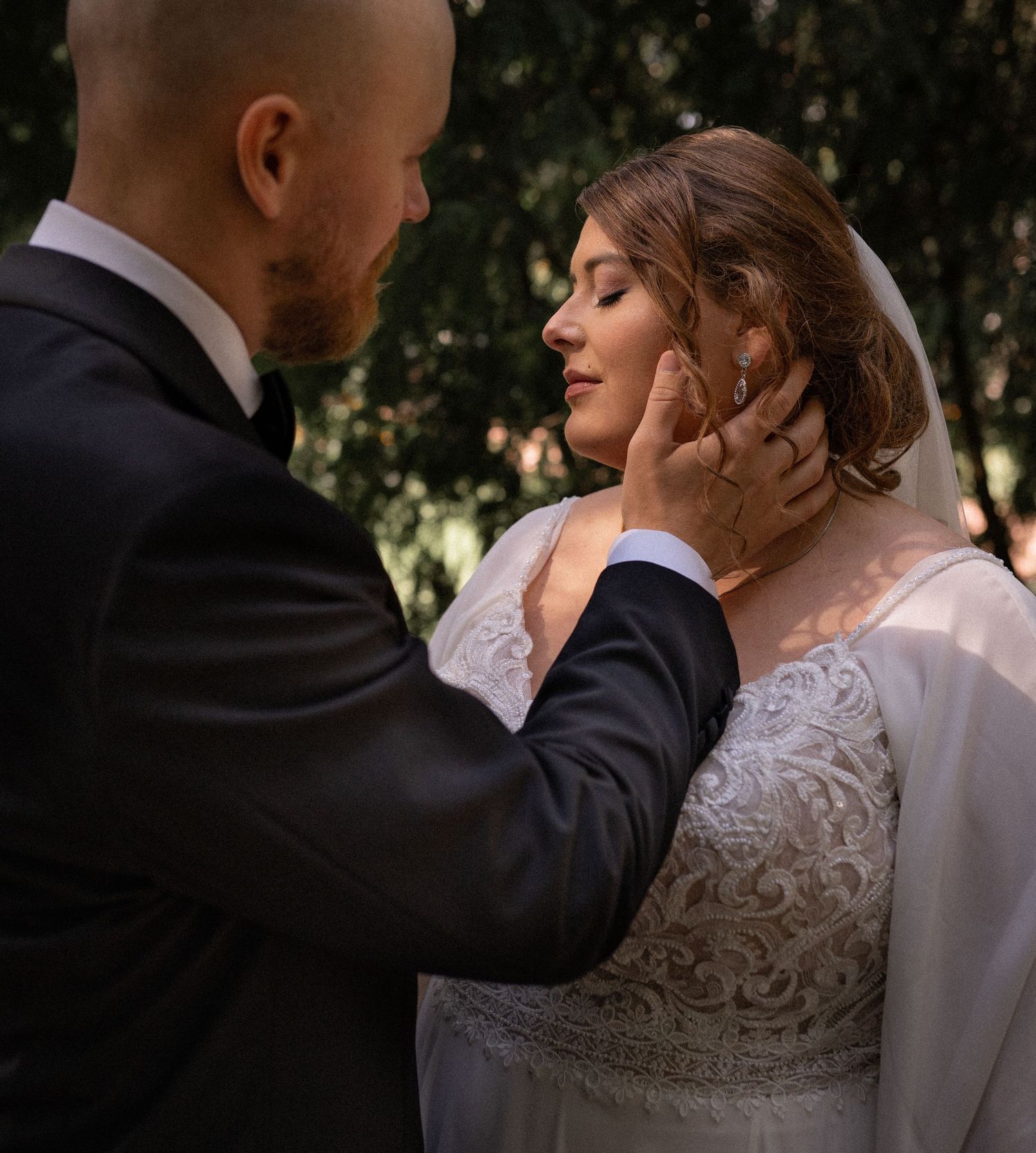 Groom gently cradles brides face as she closes her eyes in a tender outdoor wedding moment.