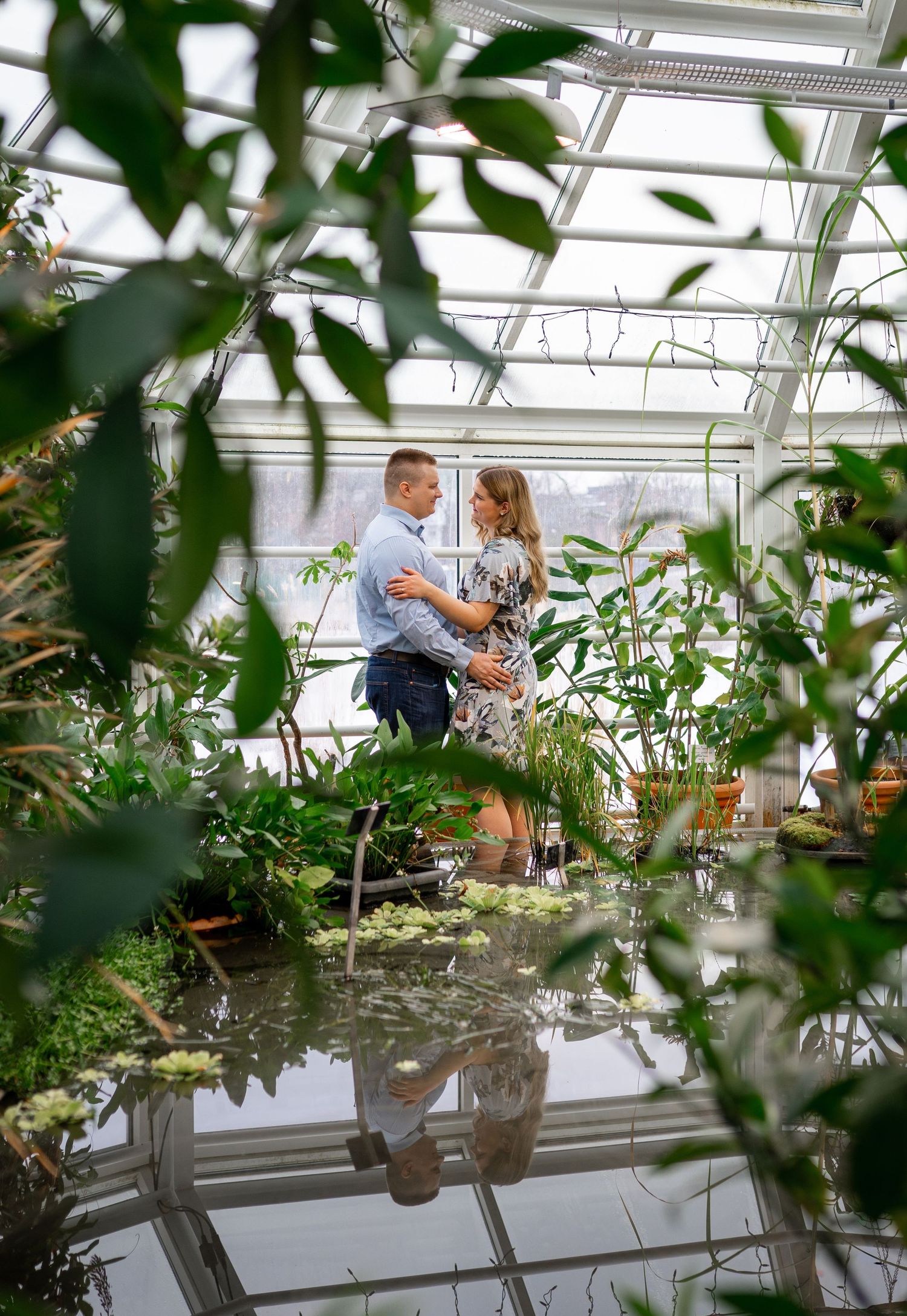 Couple embracing in a lush greenhouse surrounded by tropical plants with a reflective pool below.