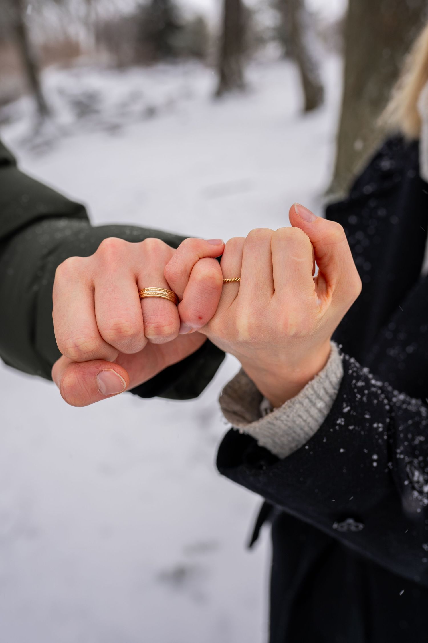 Couple linking pinky fingers outdoors in snow, both wearing gold wedding bands, romantic winter engagement photo.