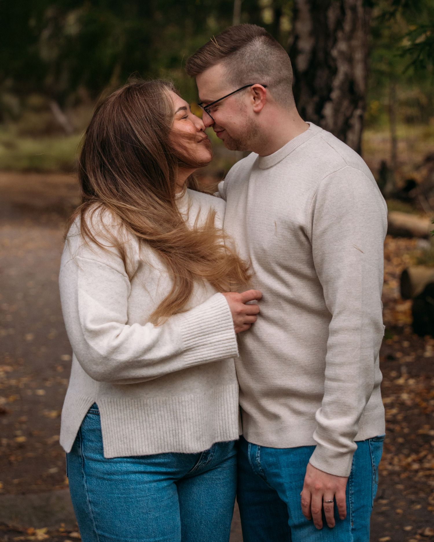 A couple in matching cream sweaters and jeans share a romantic moment on a forest path in autumn.