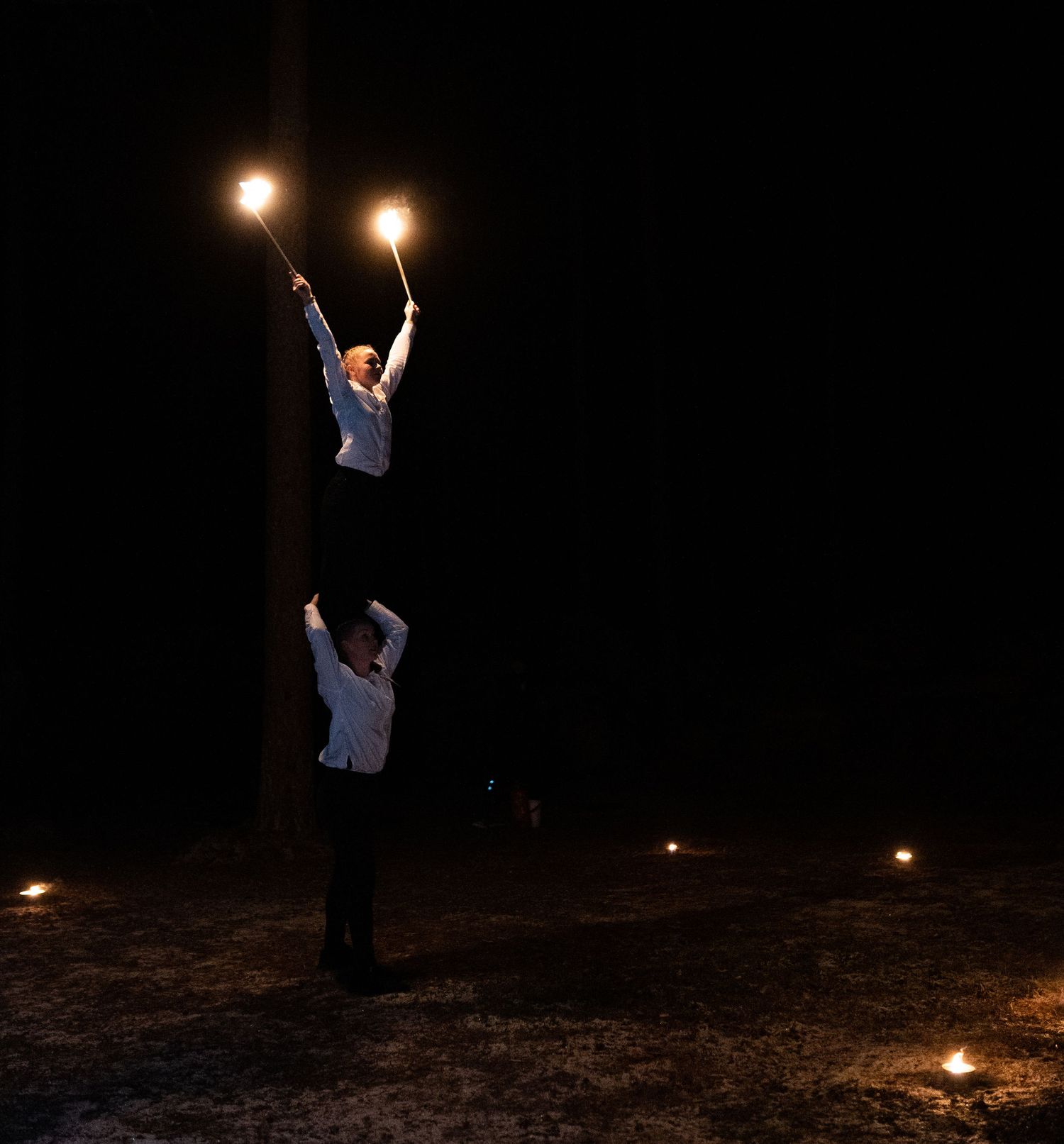 Two acrobats perform a nighttime fire juggling act, with one balancing on the other's shoulders while holding lit torches.