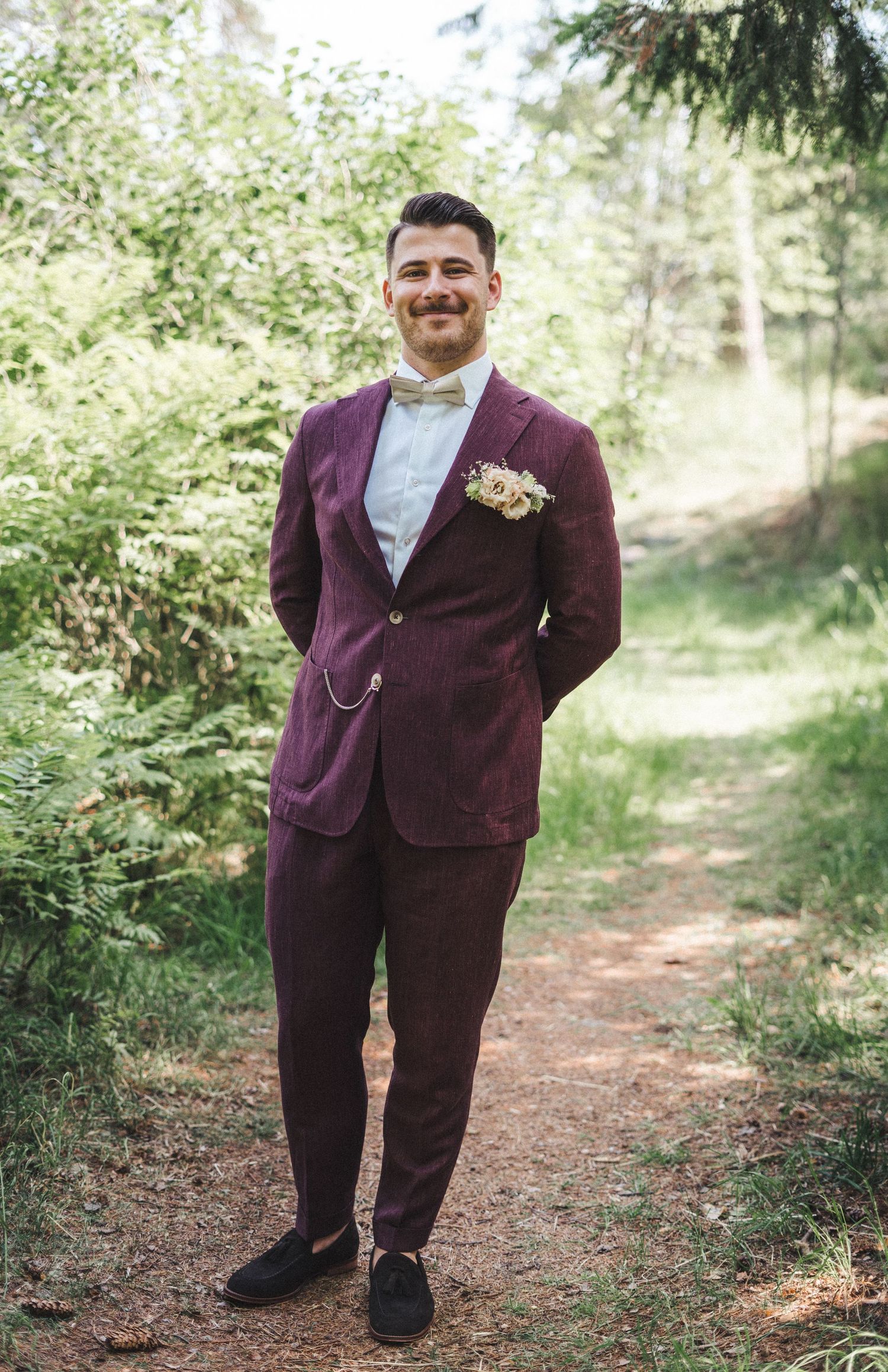 Groom in burgundy suit with bow tie and floral boutonniere standing on forest path.