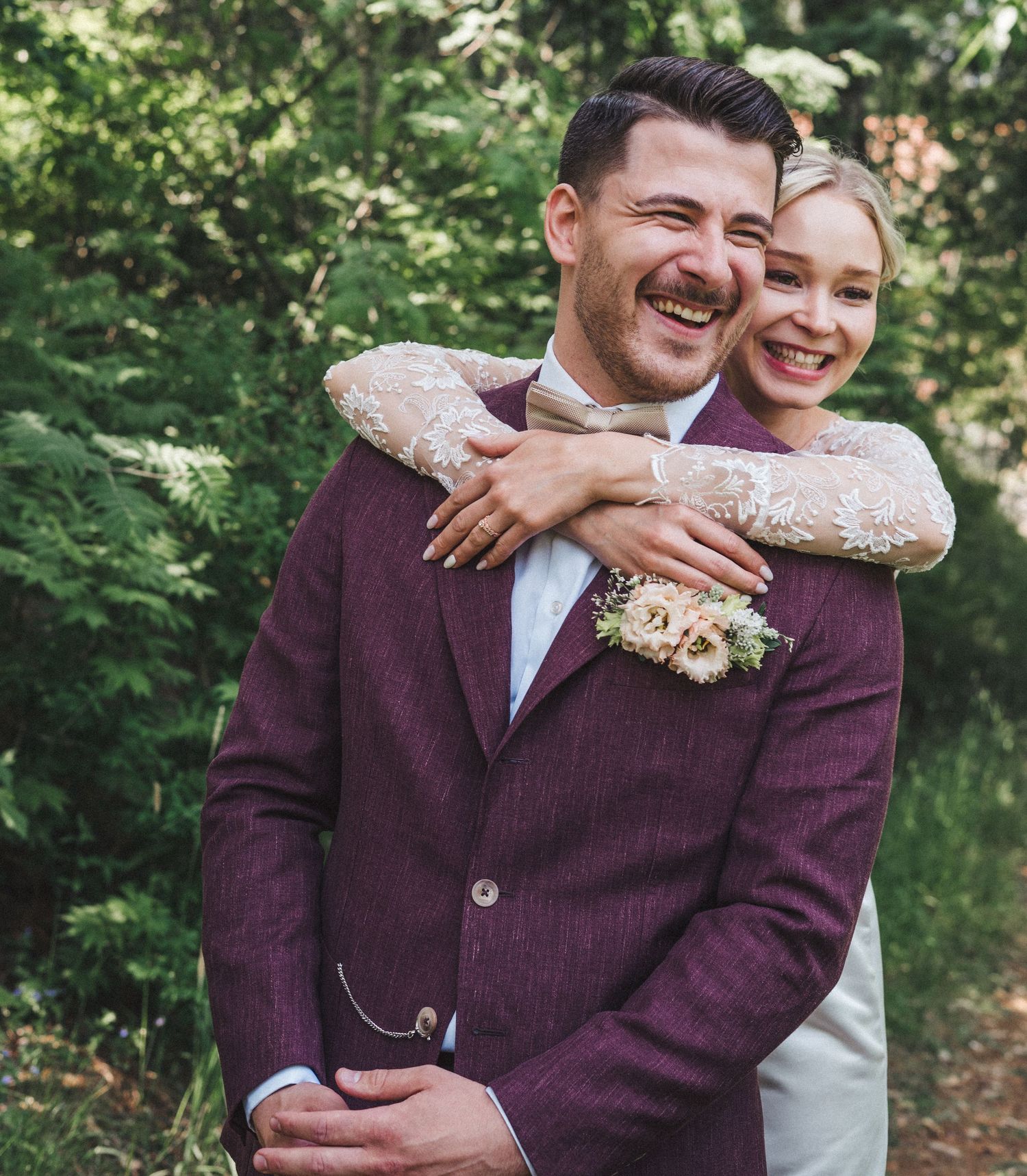 Happy bride and groom embracing outdoors, groom in burgundy suit and bride in lace wedding dress.