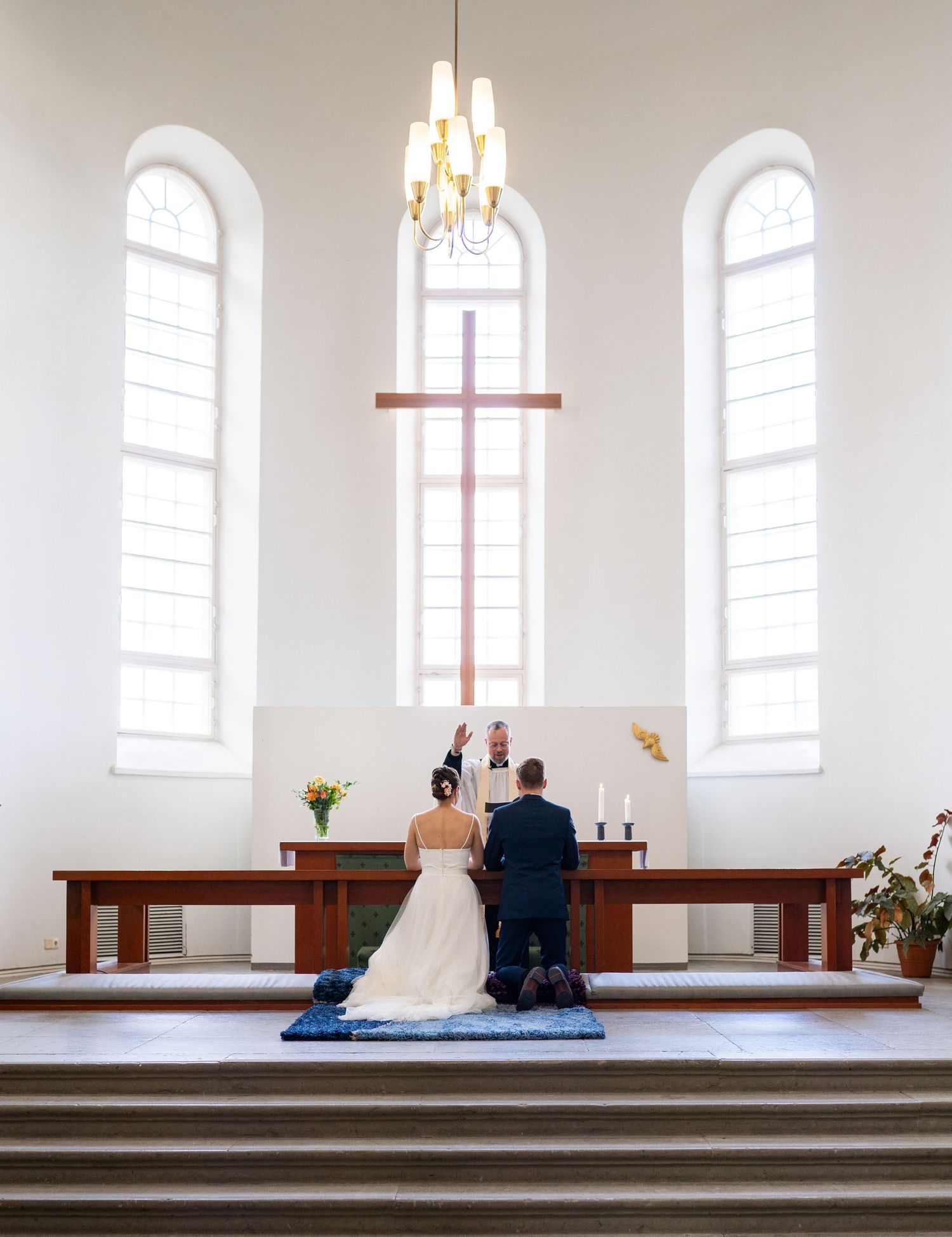 Bride and groom kneeling at church altar before large cross during wedding ceremony.