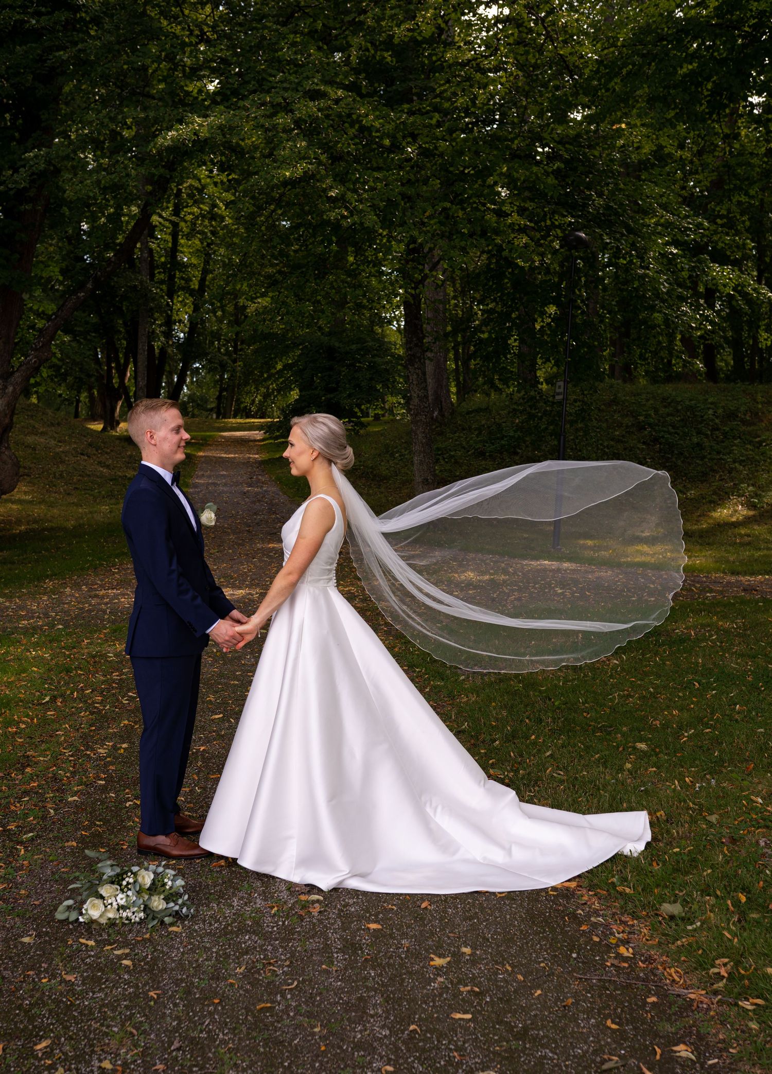 Bride and groom holding hands in tree-lined park path, brides veil flowing dramatically in the wind.
