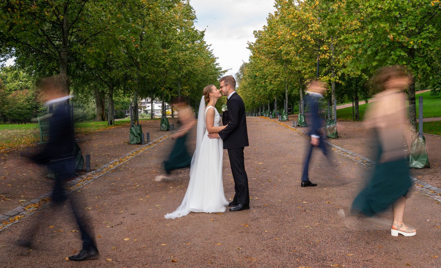 Bride and groom share a kiss as guests blur past them in a tree-lined park.