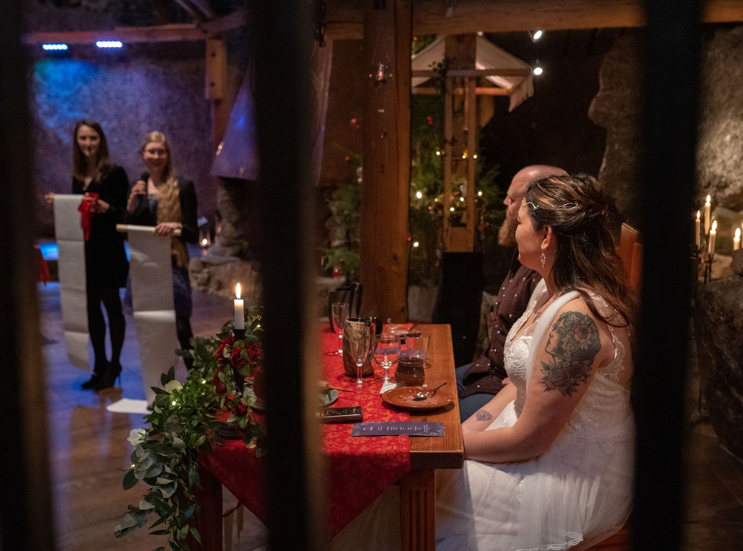 Bride in white dress sits at decorated wedding table in rustic stone venue with fairy lights and guests in background.