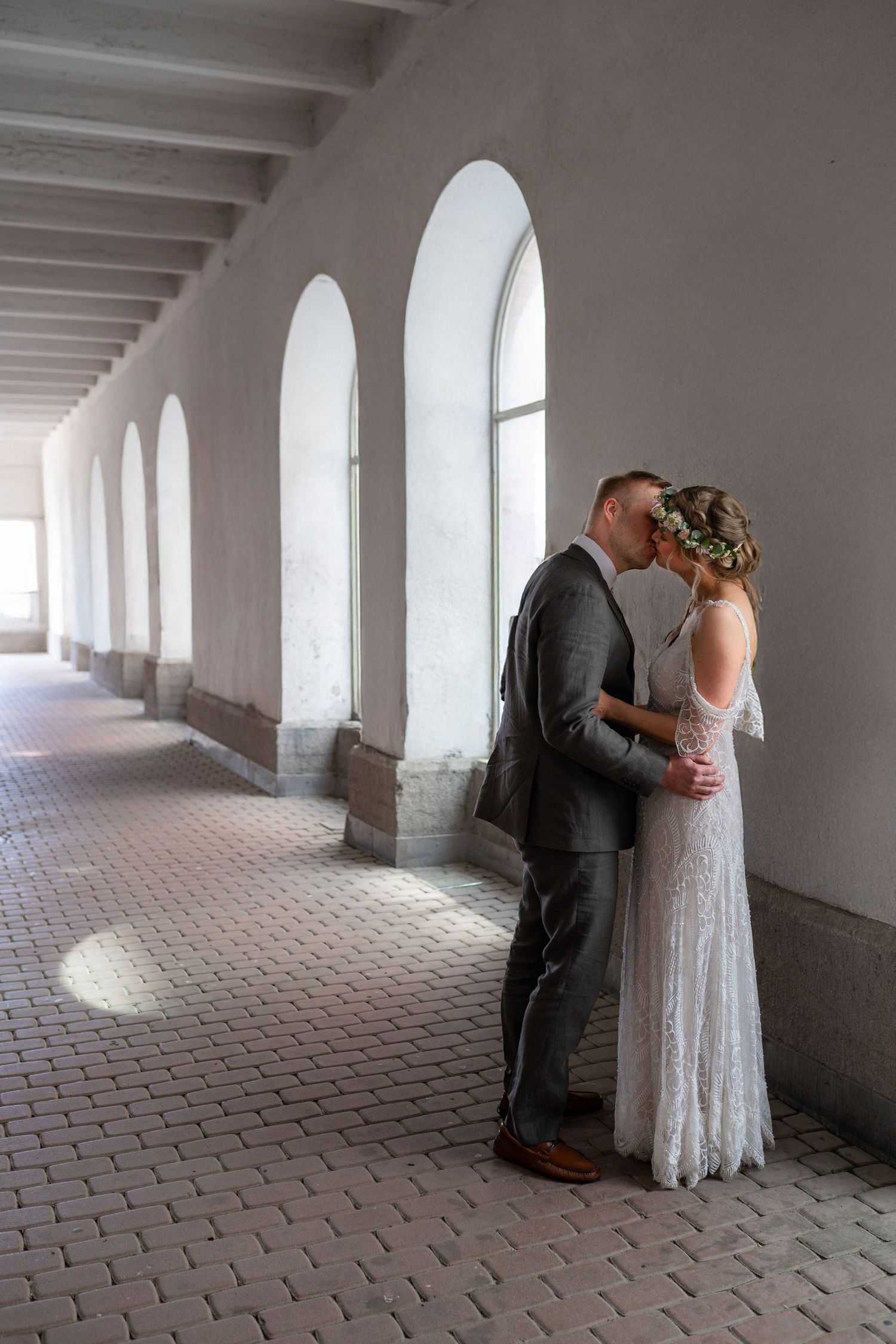 Wedding couple sharing a kiss in a sunlit arched corridor with elegant architecture.