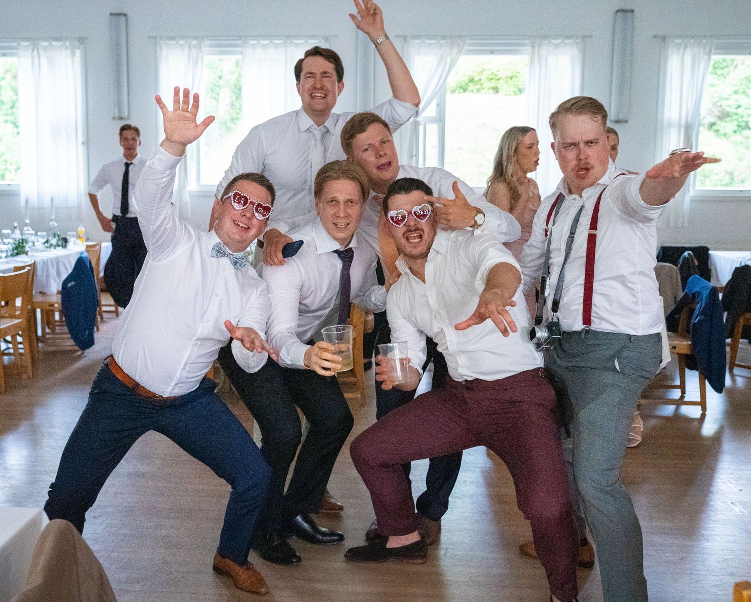 Groomsmen celebrating at a wedding reception, posing energetically in white shirts and ties in a bright venue.
