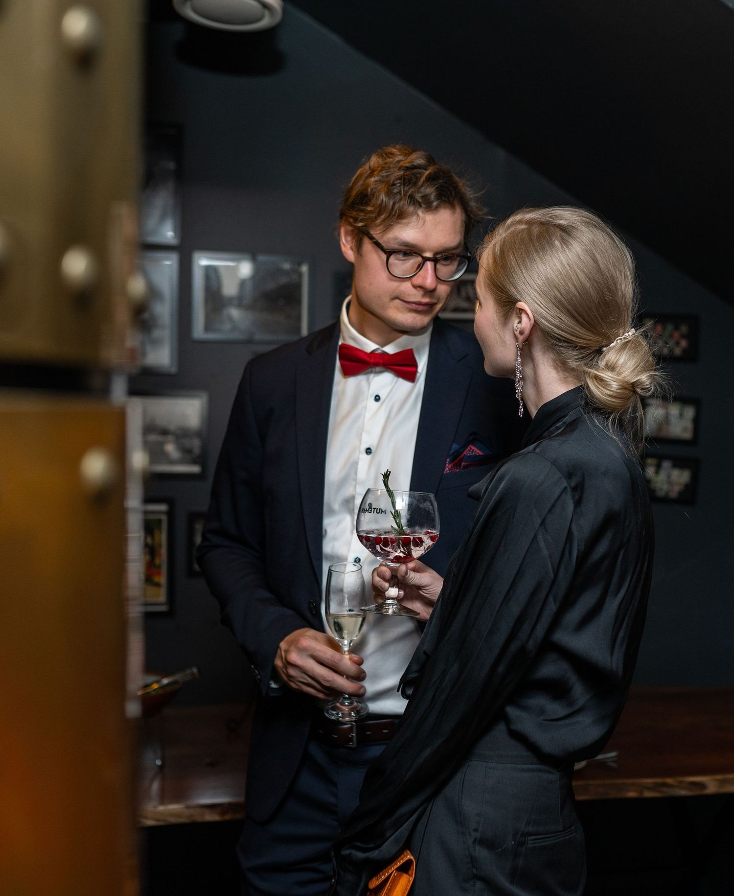 Elegant couple in formal attire sharing wine glasses at an upscale venue, man wearing navy suit with red bow tie.
