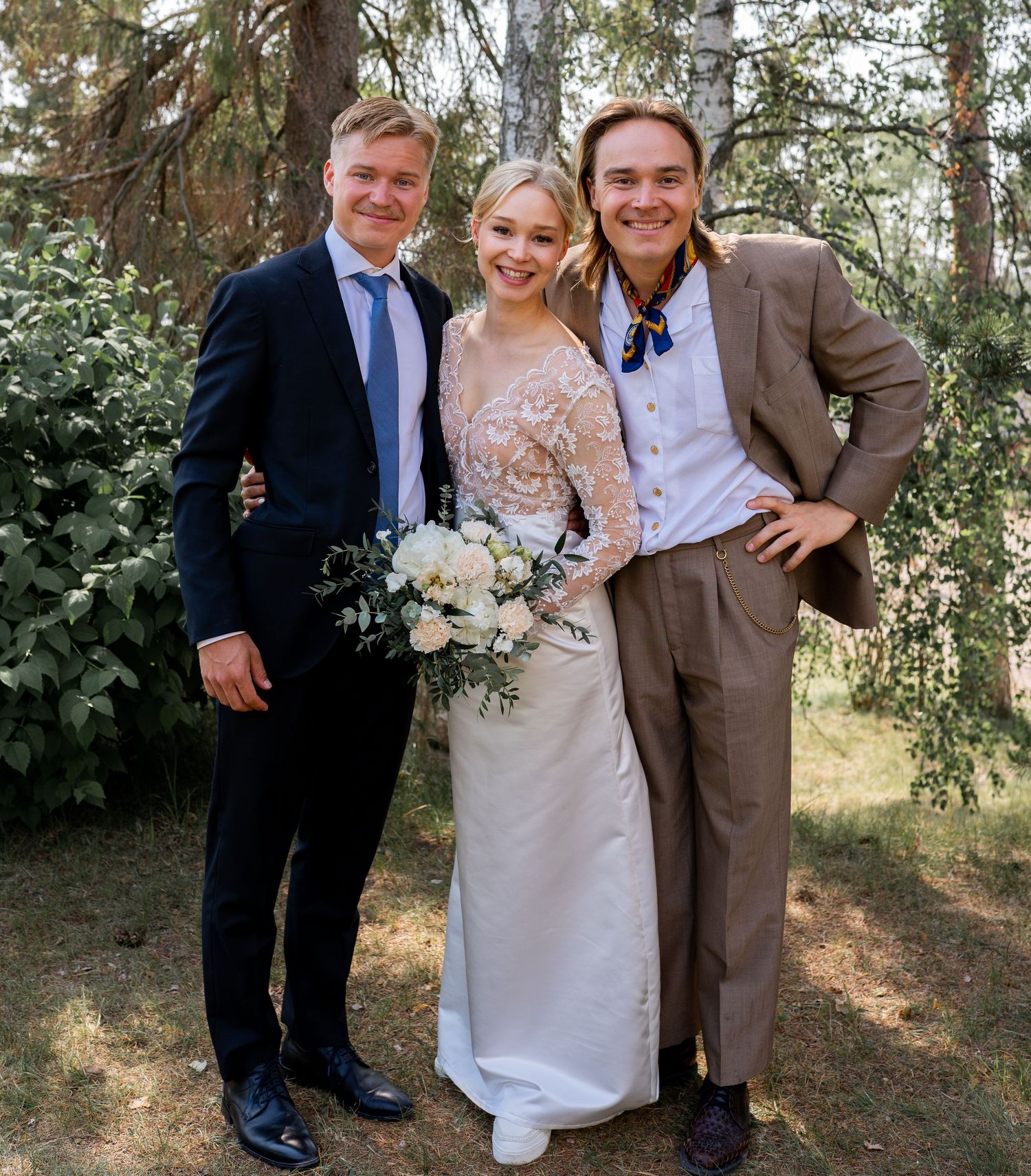 Bride holding white bouquet posing with two male wedding guests in a forest setting.