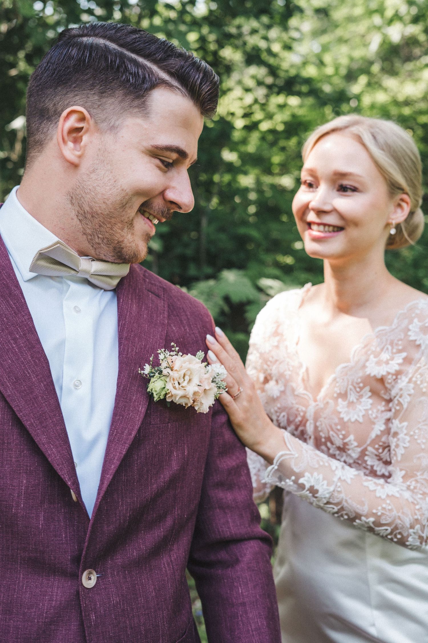 Smiling bride in lace gown touches groom's burgundy suit lapel with floral boutonniere in forest setting.
