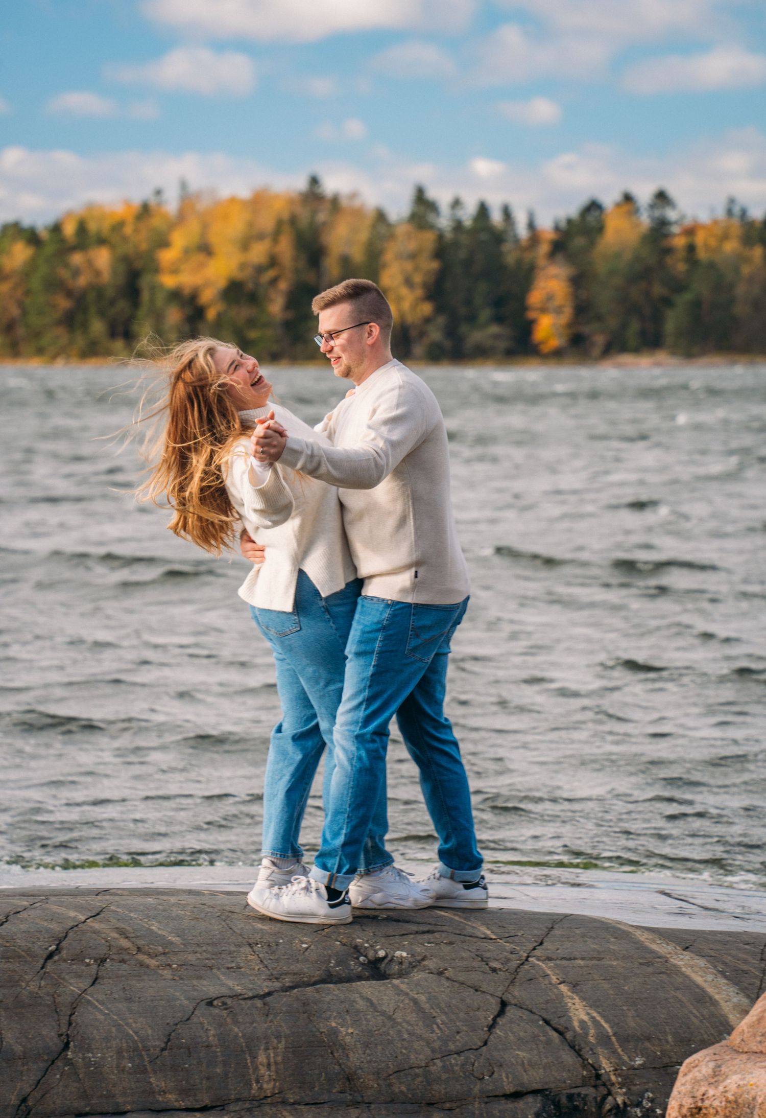A couple dances joyfully on rocky shore by a lake, wearing white jackets and blue jeans, with autumn trees in background.