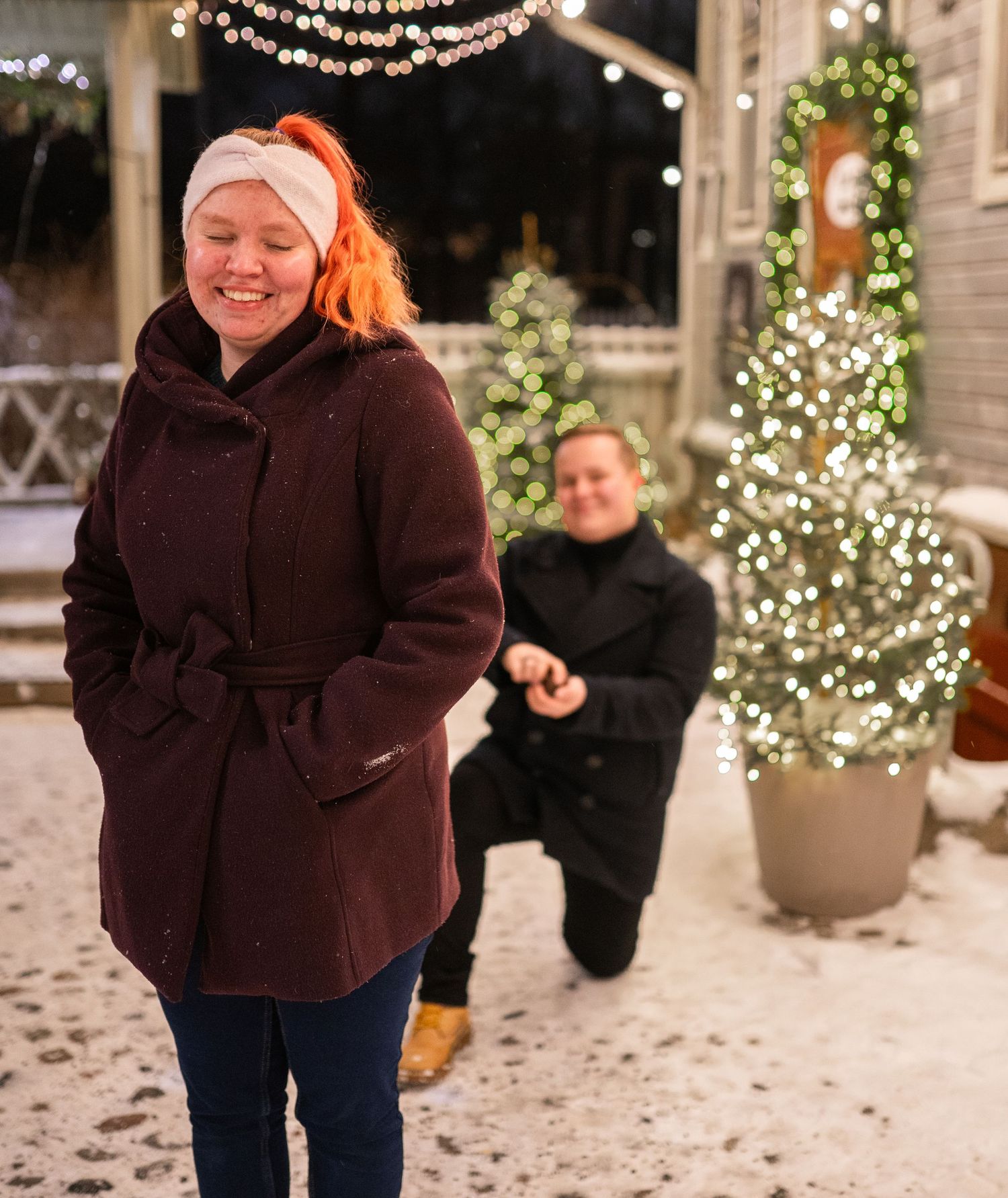 Man proposing on snowy winter night with Christmas lights while woman smiles joyfully in maroon coat with orange hair.