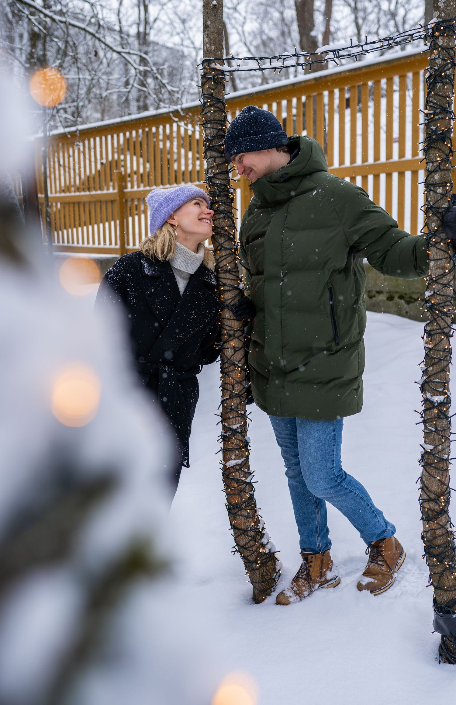 Couple smiling at each other outdoors in snowfall, wearing winter coats and hats near tree-wrapped lights.