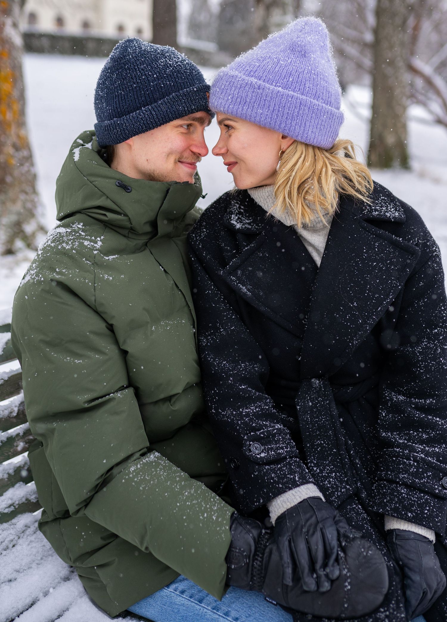 Couple in winter hats and coats sitting close on a snowy bench, sharing a romantic moment outdoors.