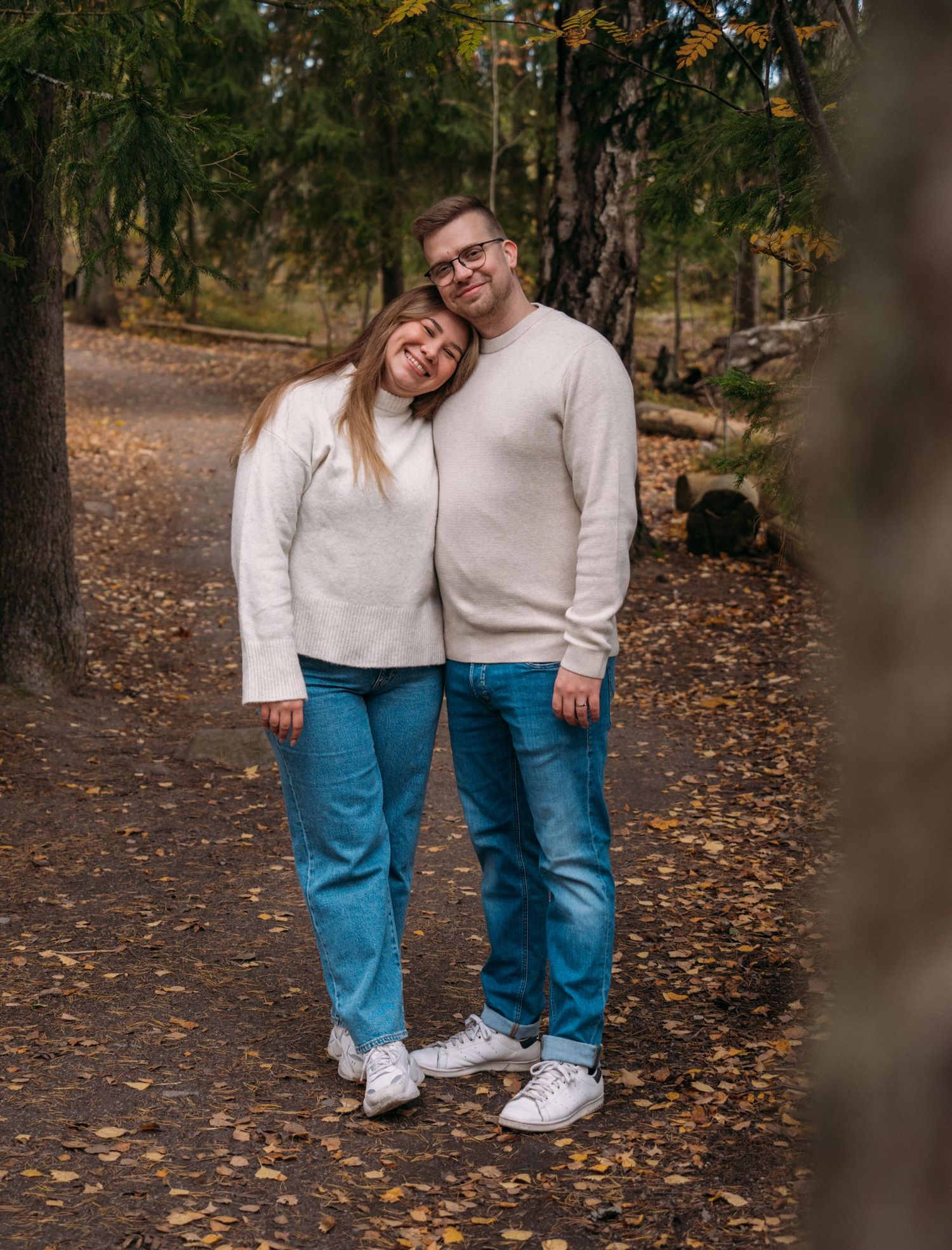Couple in matching white sweaters and blue jeans posing together on an autumn forest path covered in fallen leaves.