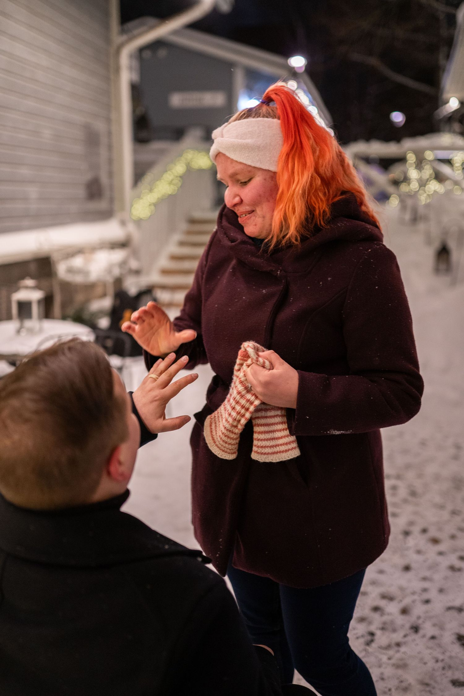 Man kneeling in snow proposing to red-haired woman in burgundy coat during winter night outdoors.