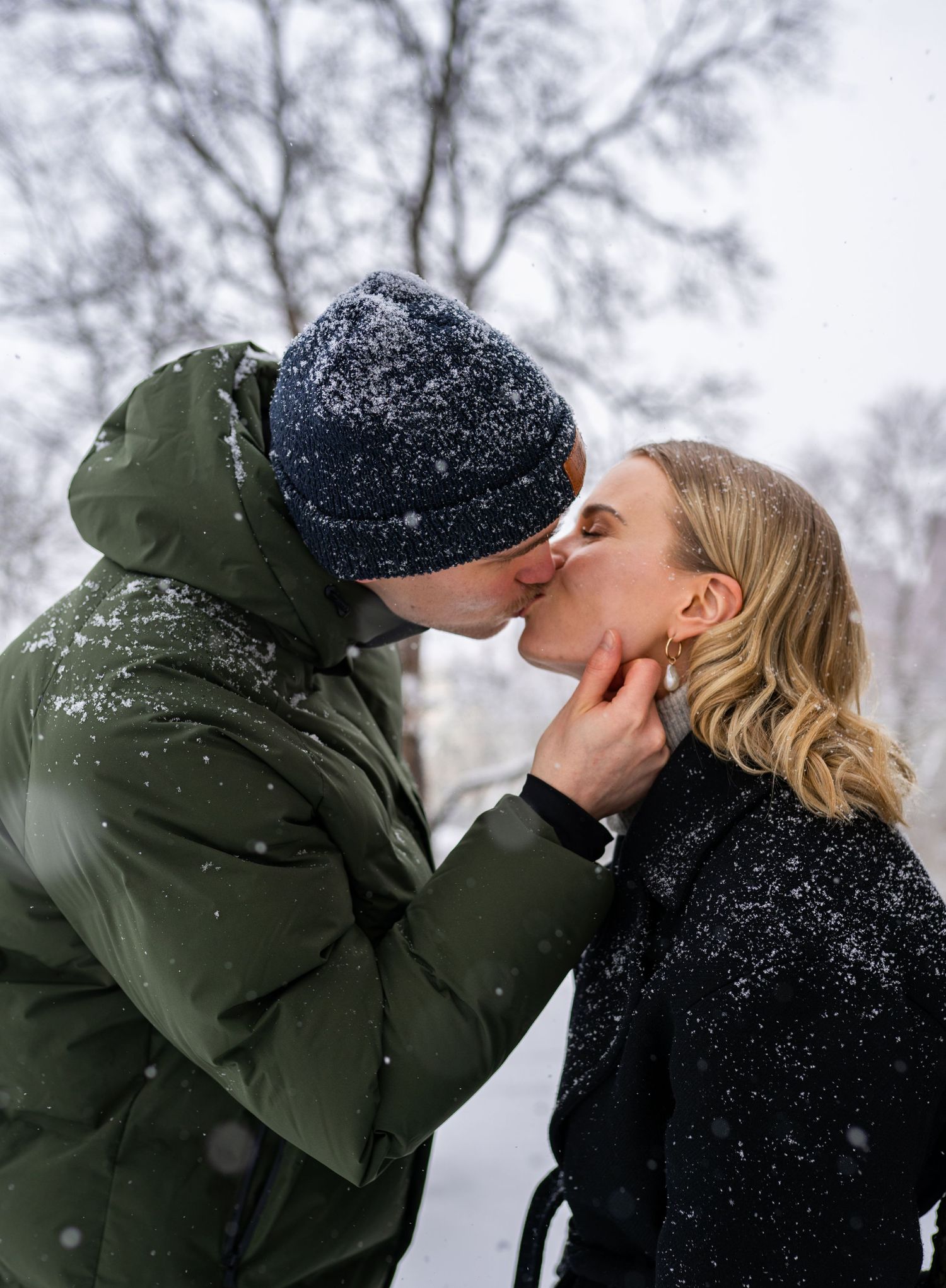 Couple sharing a romantic kiss in falling snow, wearing winter coats, with snow-covered trees in the background.