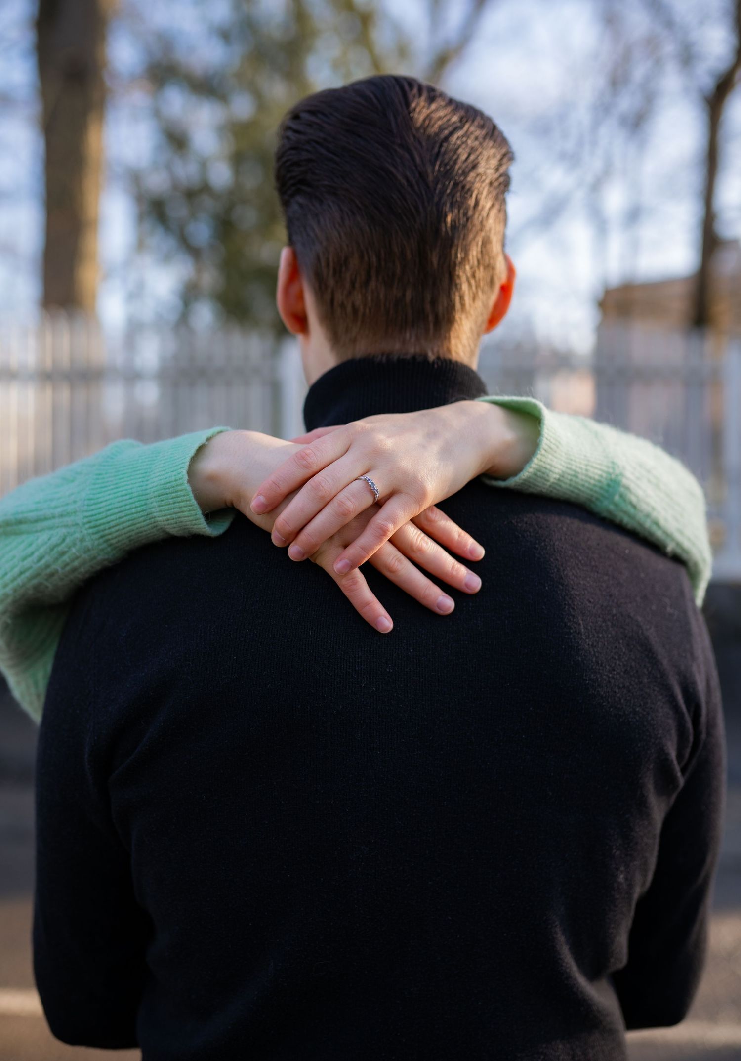 A person in a green sweater rests their hand on the shoulder of someone in a black sweater, showing an engagement ring.