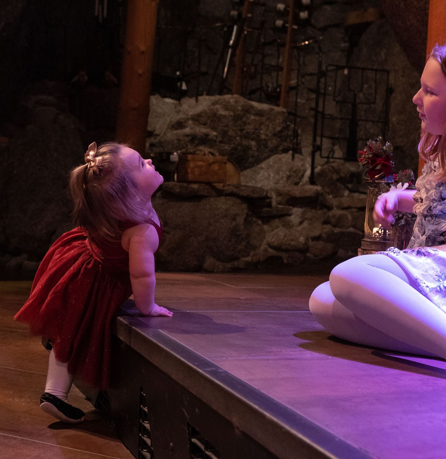 Two young girls interact on a stage, one in a red dress standing below and one in white sitting above her.