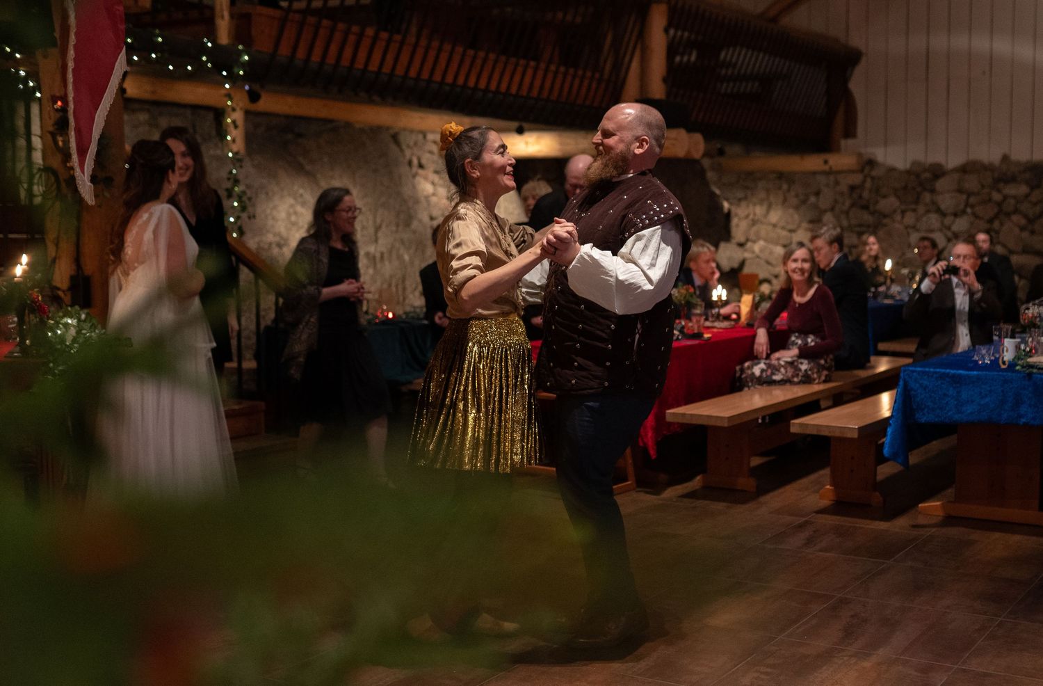Couple dancing in medieval costumes at a festive barn celebration with guests seated at long wooden tables.