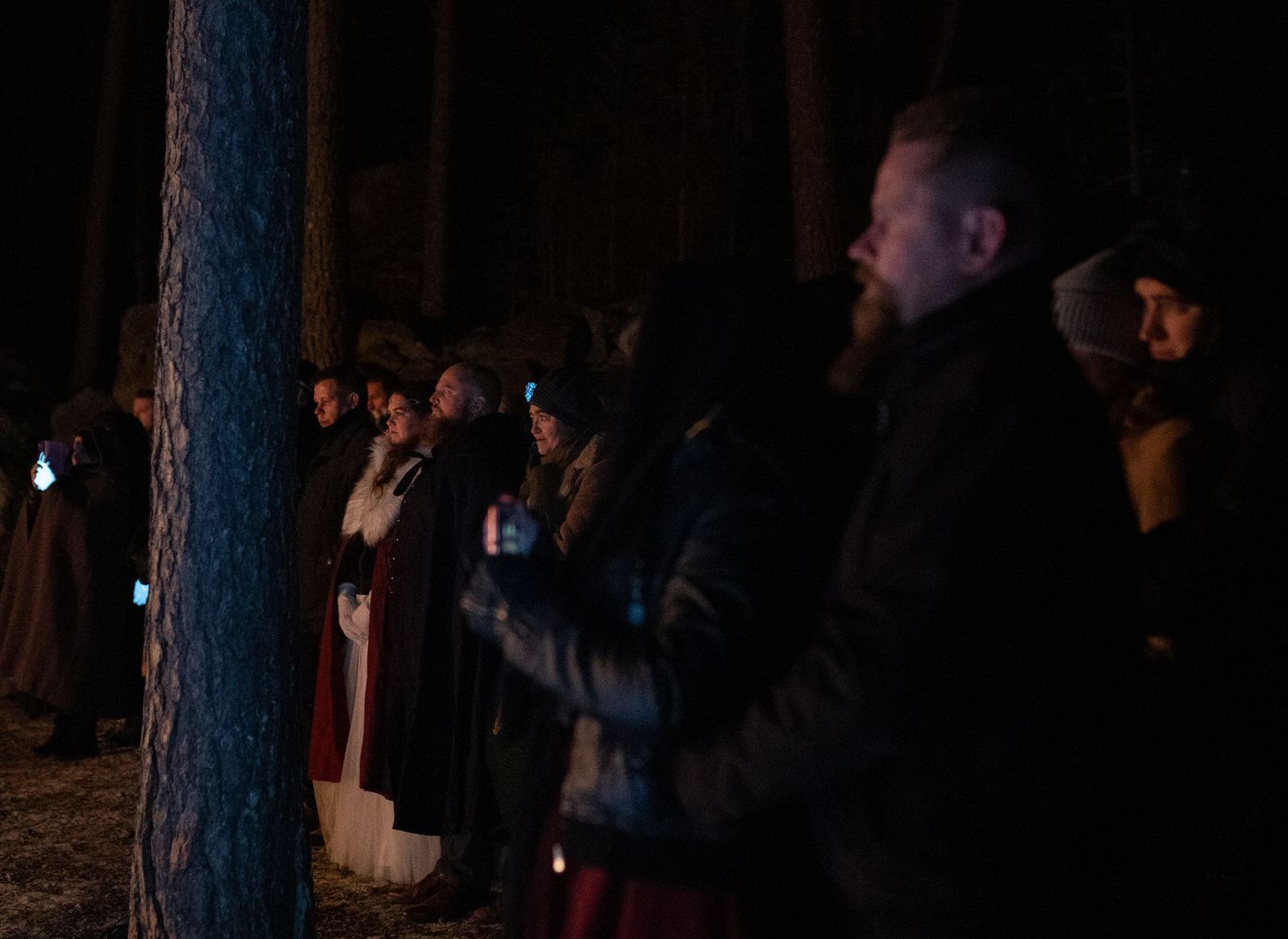 Group of people standing in a dark forest at night, holding candles during an outdoor ceremony.