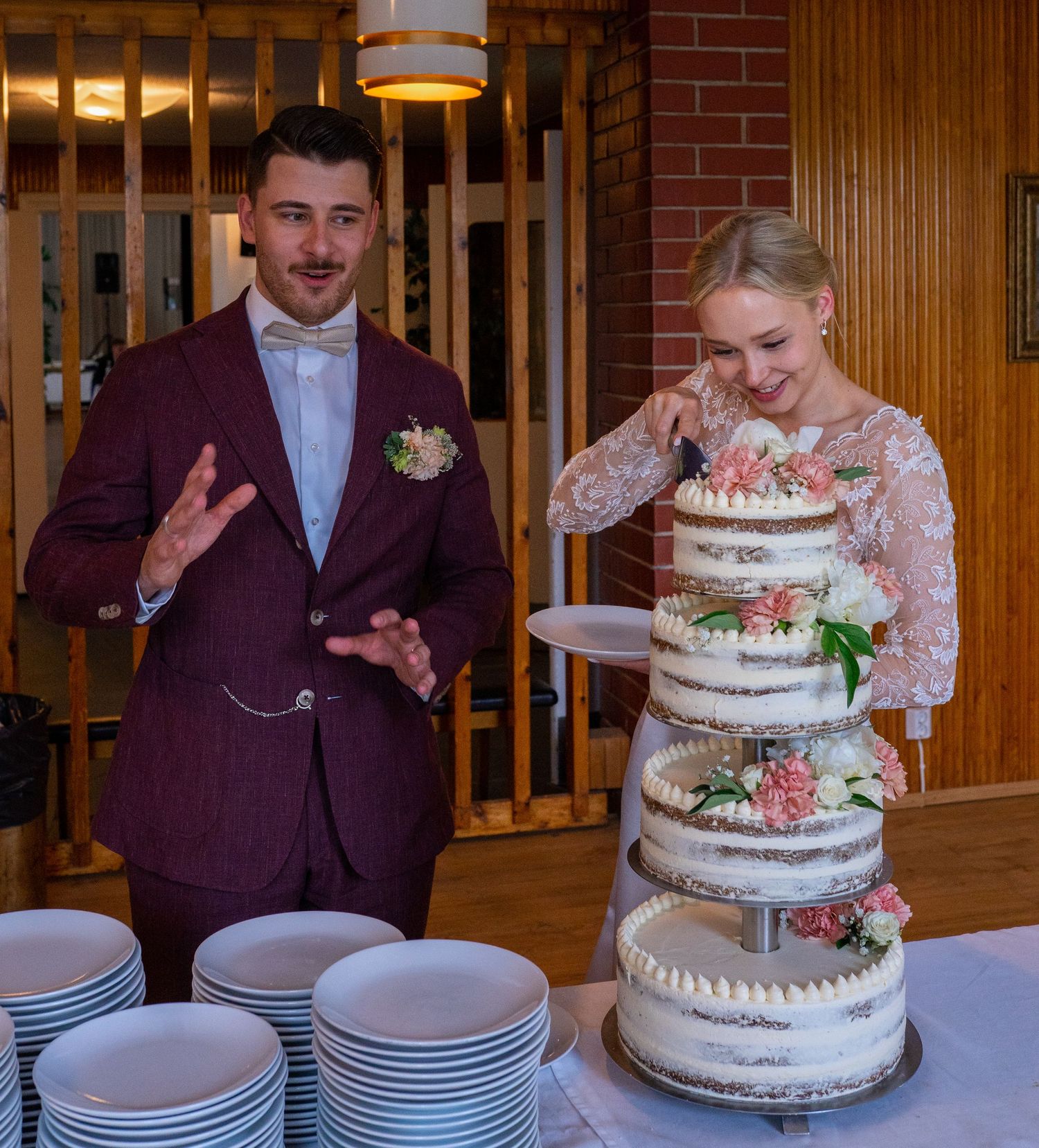 Bride laughs while groom gestures proudly beside their elegant three-tier floral naked wedding cake.