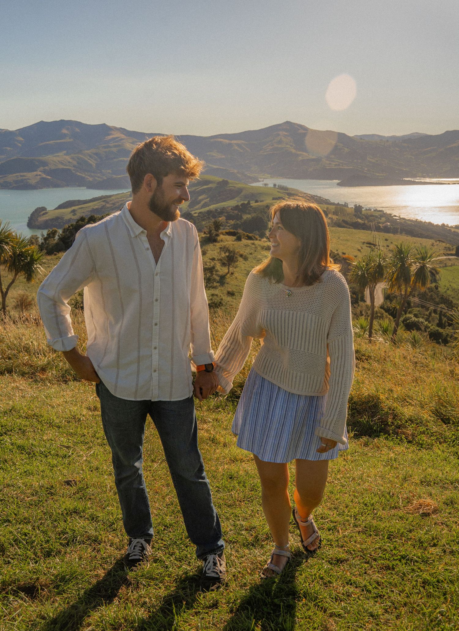 Couple holding hands on a scenic hilltop overlooking a bay and mountains at golden hour.