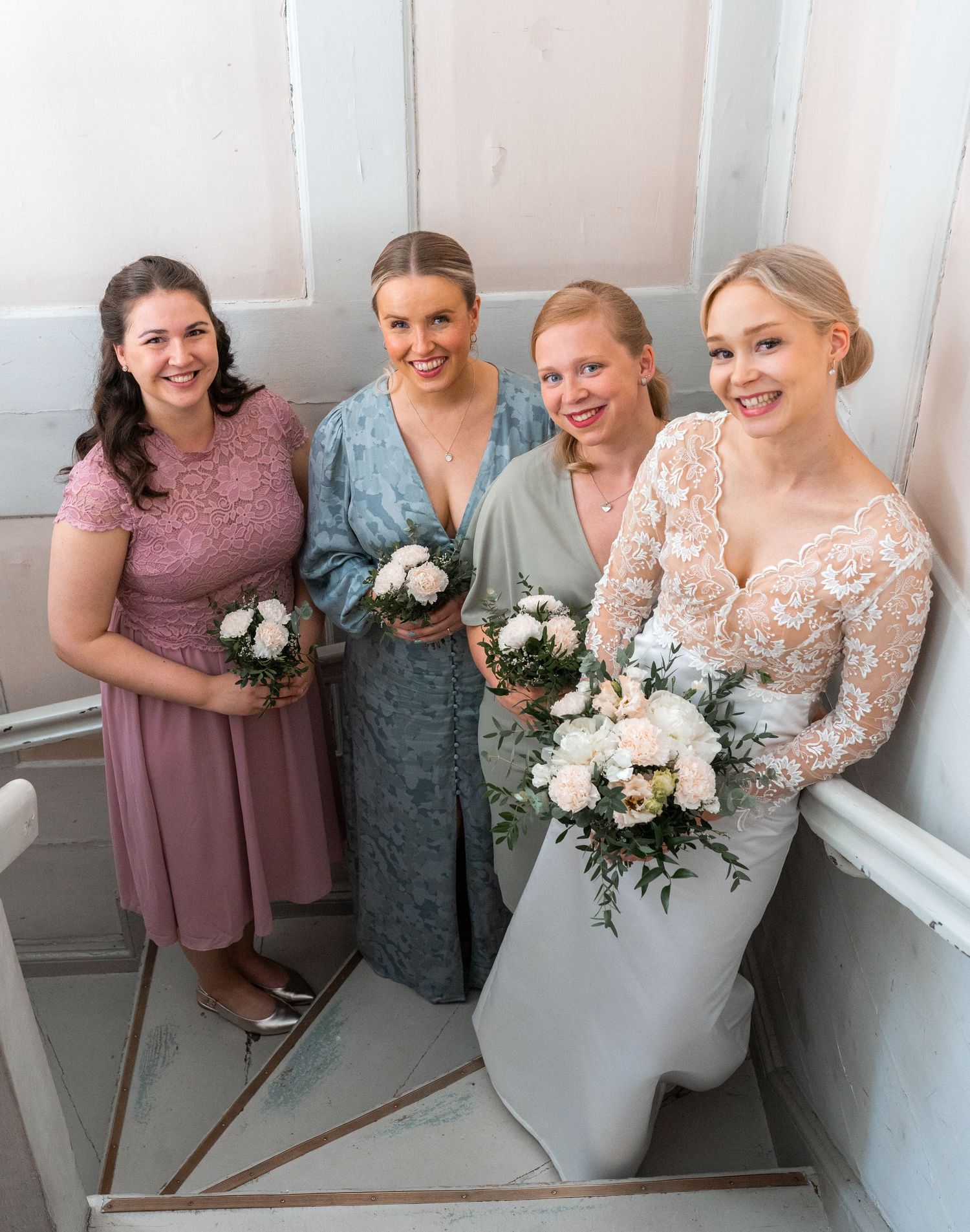 Bride in lace gown with three bridesmaids in pink and blue dresses holding bouquets on stairs.