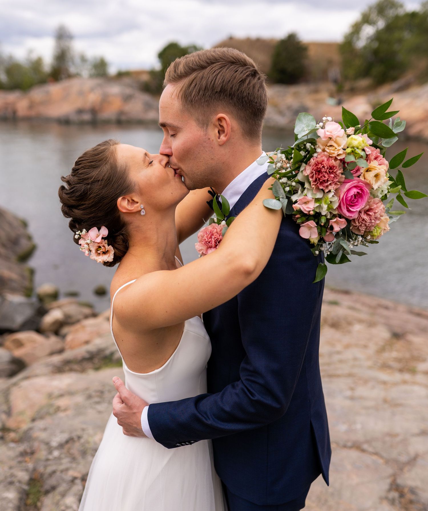 Bride and groom sharing a kiss outdoors on rocky shore, bride holding pink bouquet.