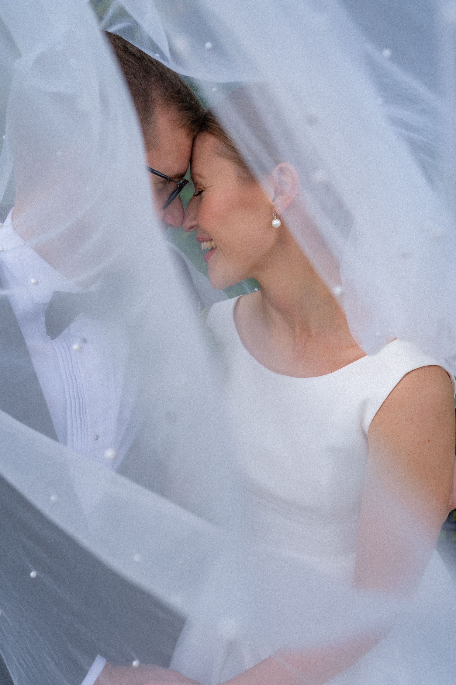 Intimate wedding couple touching foreheads tenderly beneath a flowing white veil.
