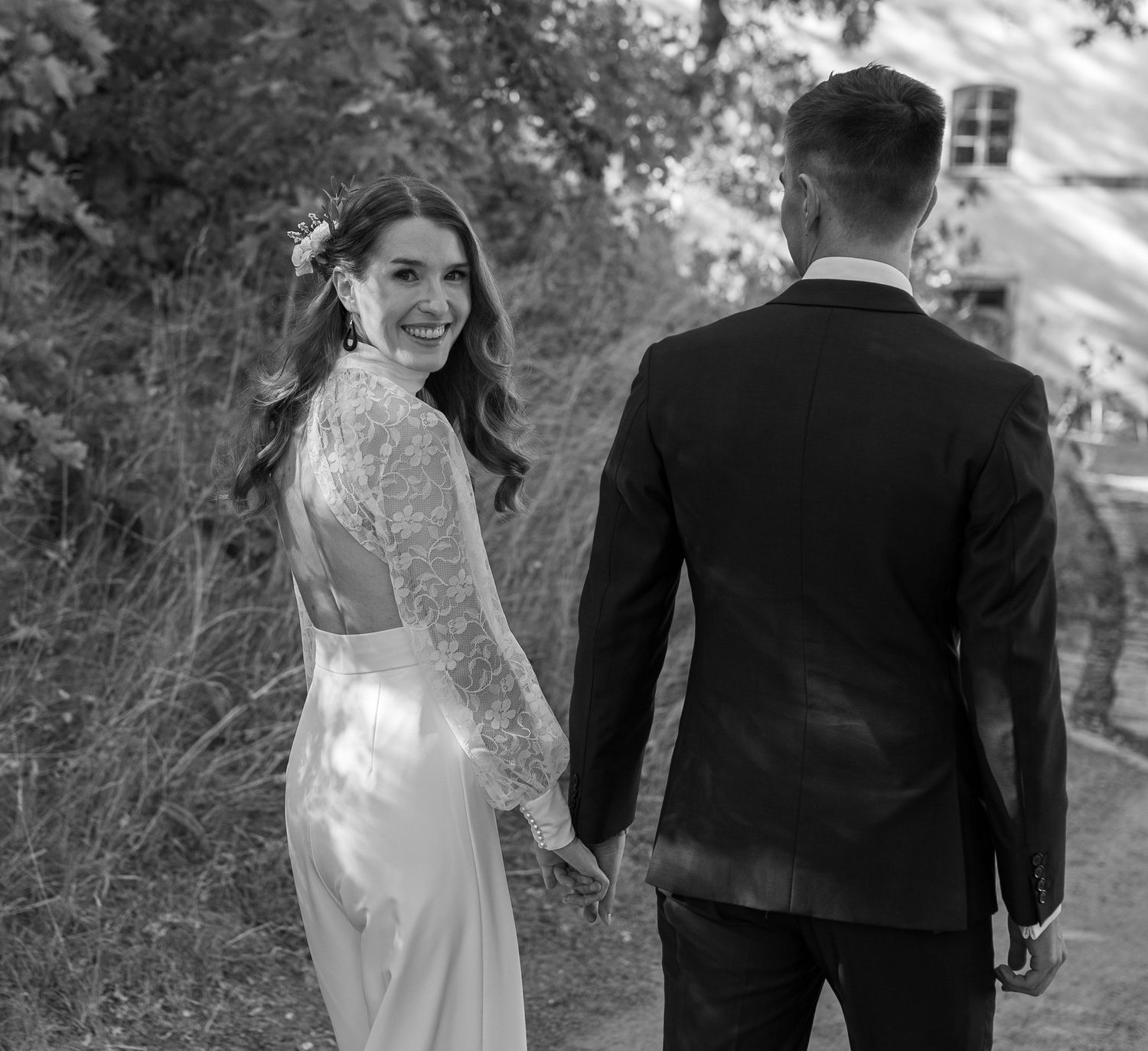 Black and white photo of smiling bride in lace gown holding grooms hand on path.