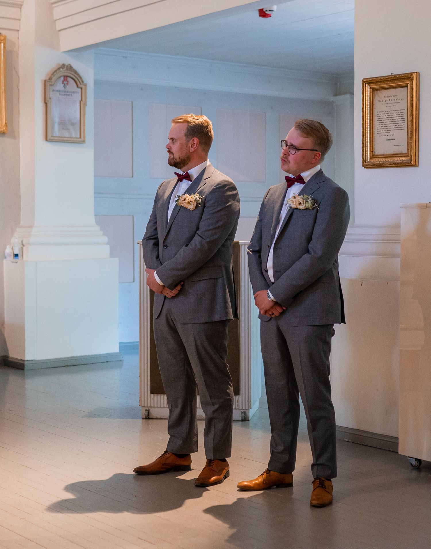 Two groomsmen in matching grey suits with bow ties and boutonnieres stand attentively during wedding ceremony.