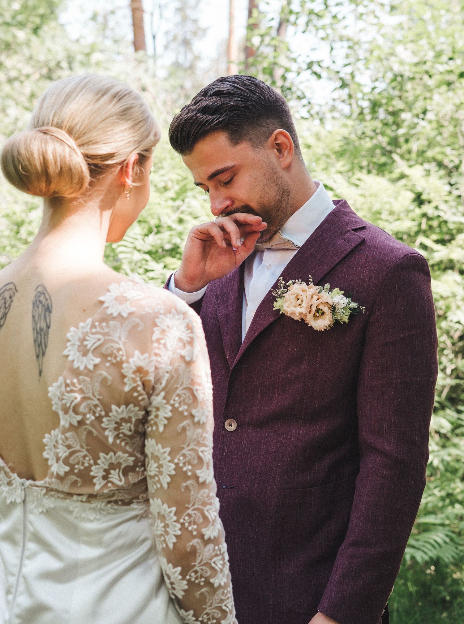 Groom in purple tweed suit wipes tear during intimate first look moment with bride in lace wedding dress.