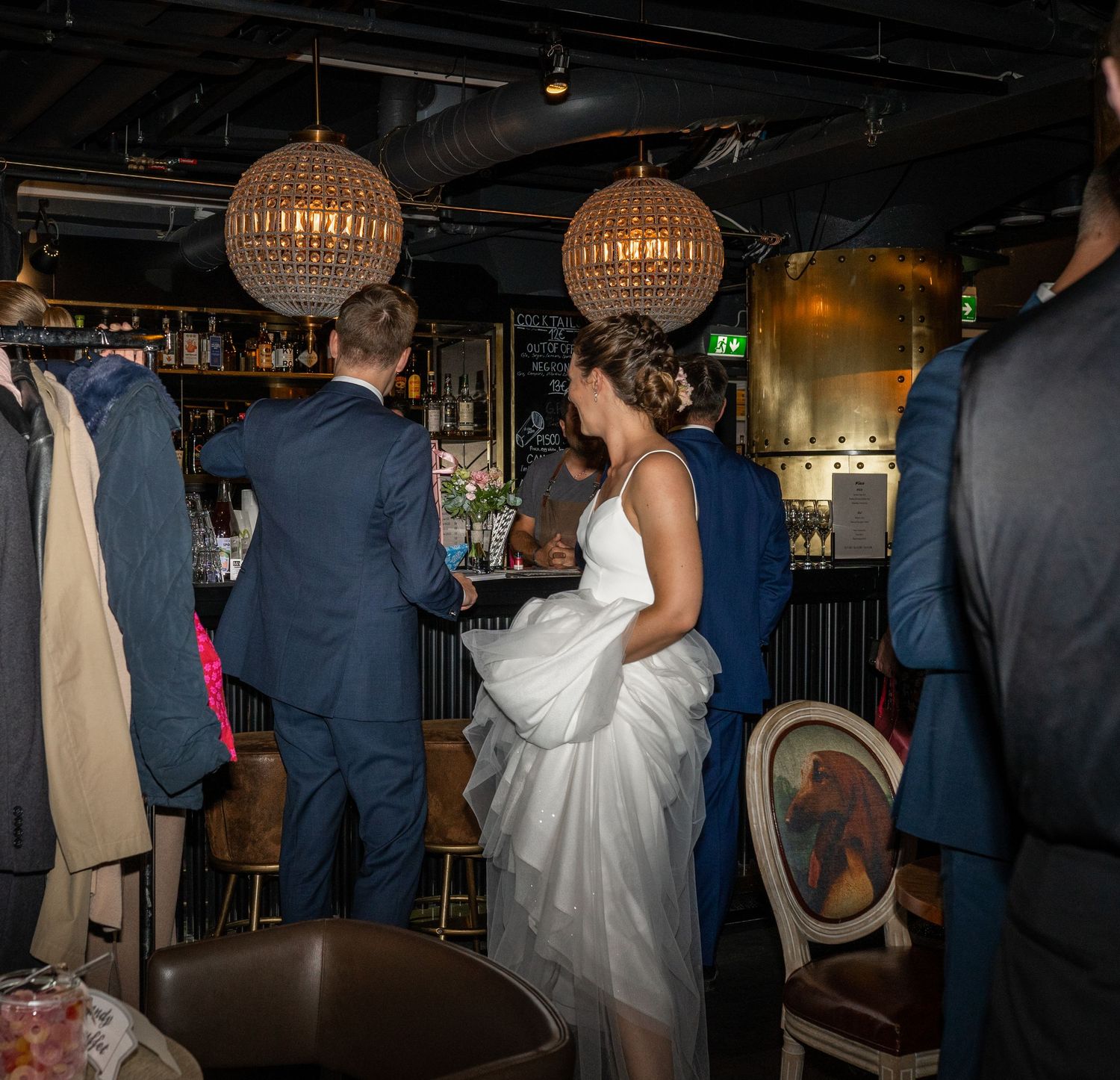 Bride and groom stand together at bar in dimly lit wedding reception venue with pendant lights.