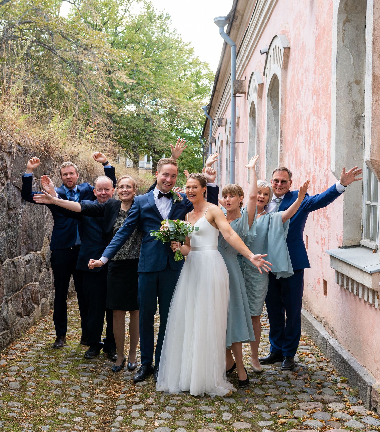 Joyful wedding party of eight celebrates with raised arms on cobblestone path beside pink building.