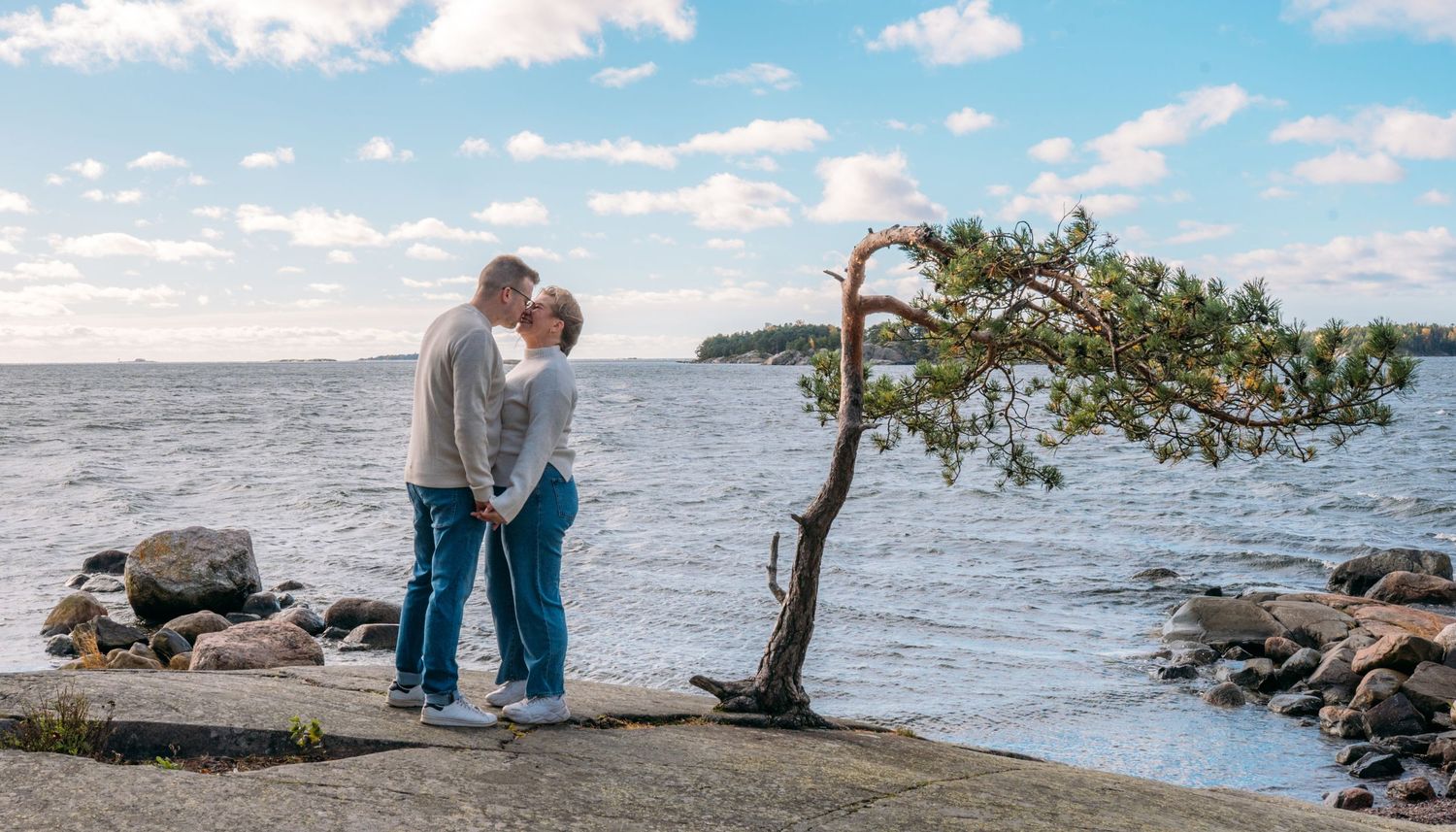 Couple holding hands and sharing a romantic moment by the rocky shoreline near a windswept pine tree on a cloudy day.