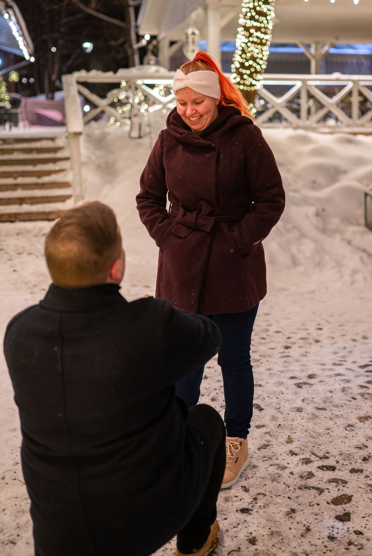 Man kneeling in snow proposing to smiling woman in burgundy coat and red hat amid festive Christmas lights.