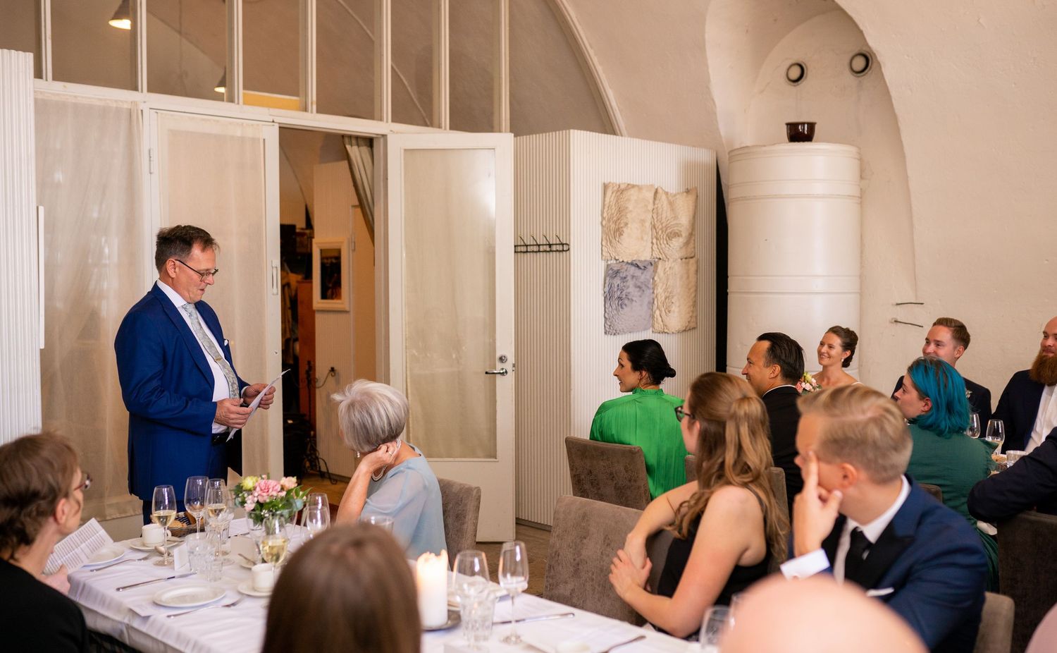 Wedding speaker in blue suit addressing seated guests at elegant reception dinner with arched ceilings.