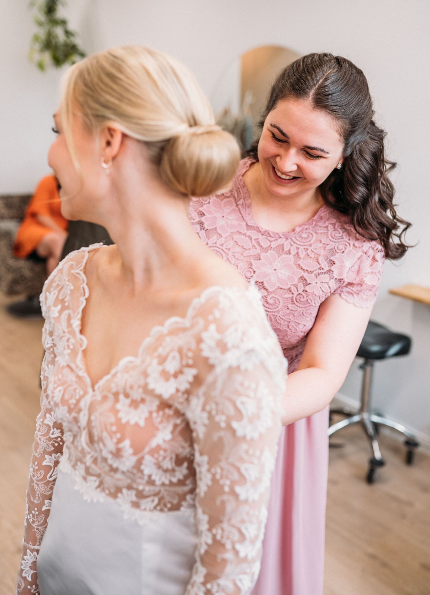Smiling bridesmaid in pink lace dress helping a blonde bride with her lace wedding gown.