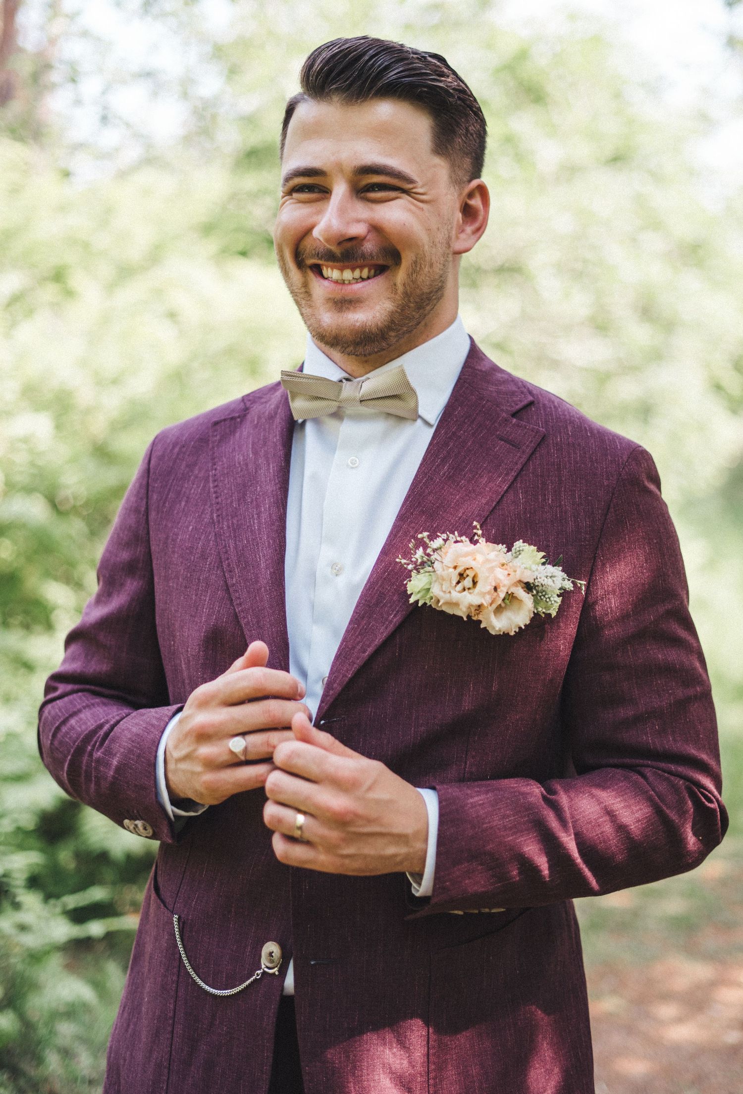 Smiling groom in burgundy suit with beige bow tie and floral boutonniere outdoors.