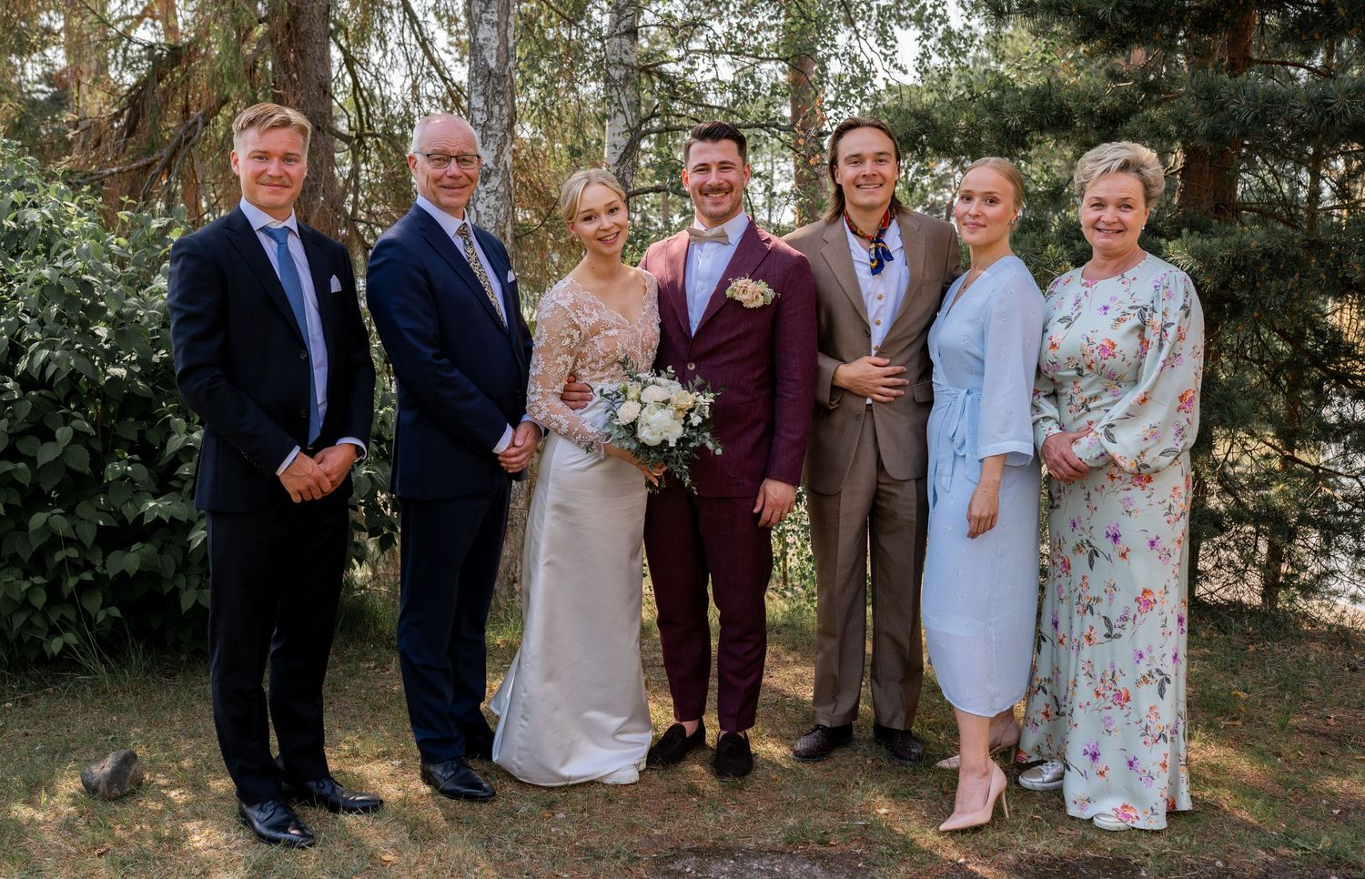Wedding party group photo with bride, groom, and family members outdoors among pine trees.