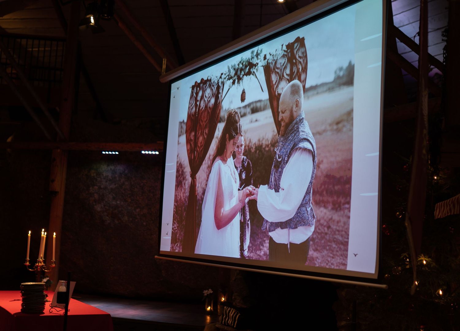 Wedding photo displayed on projector screen in rustic venue with candles, showing couple exchanging rings outdoors.