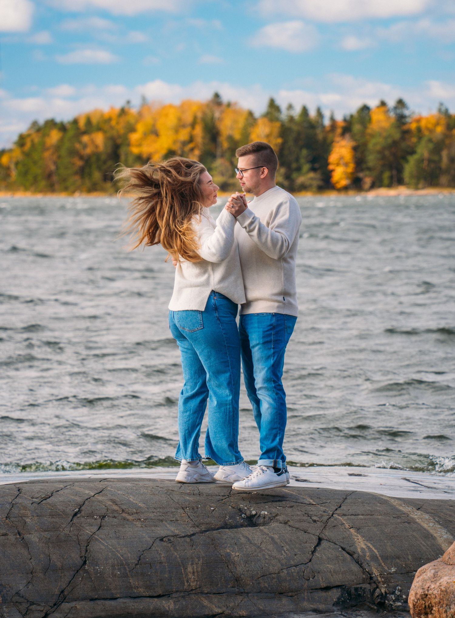 Couple in matching white sweaters and jeans dancing on rocks by a lake with autumn foliage in background.