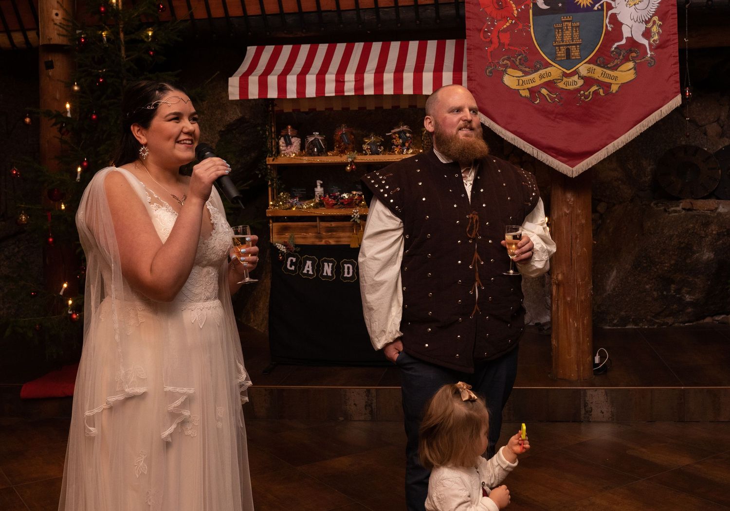Couple in medieval costumes laughing at a Renaissance faire wedding reception with a child nearby.
