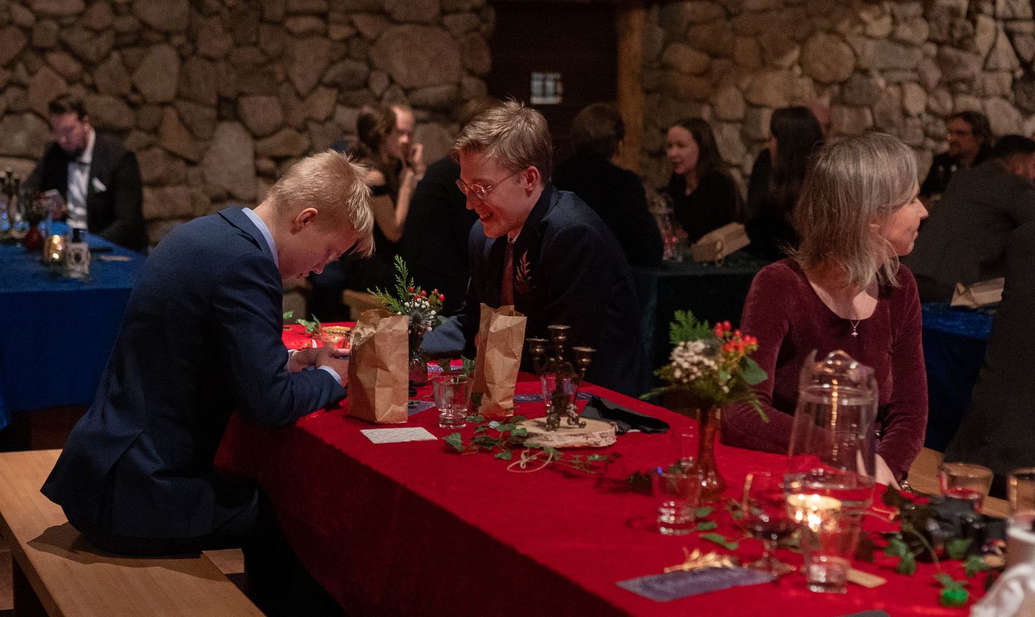 Guests mingle at an elegant holiday dinner event with red tablecloths, candles, and festive decor in a rustic venue.
